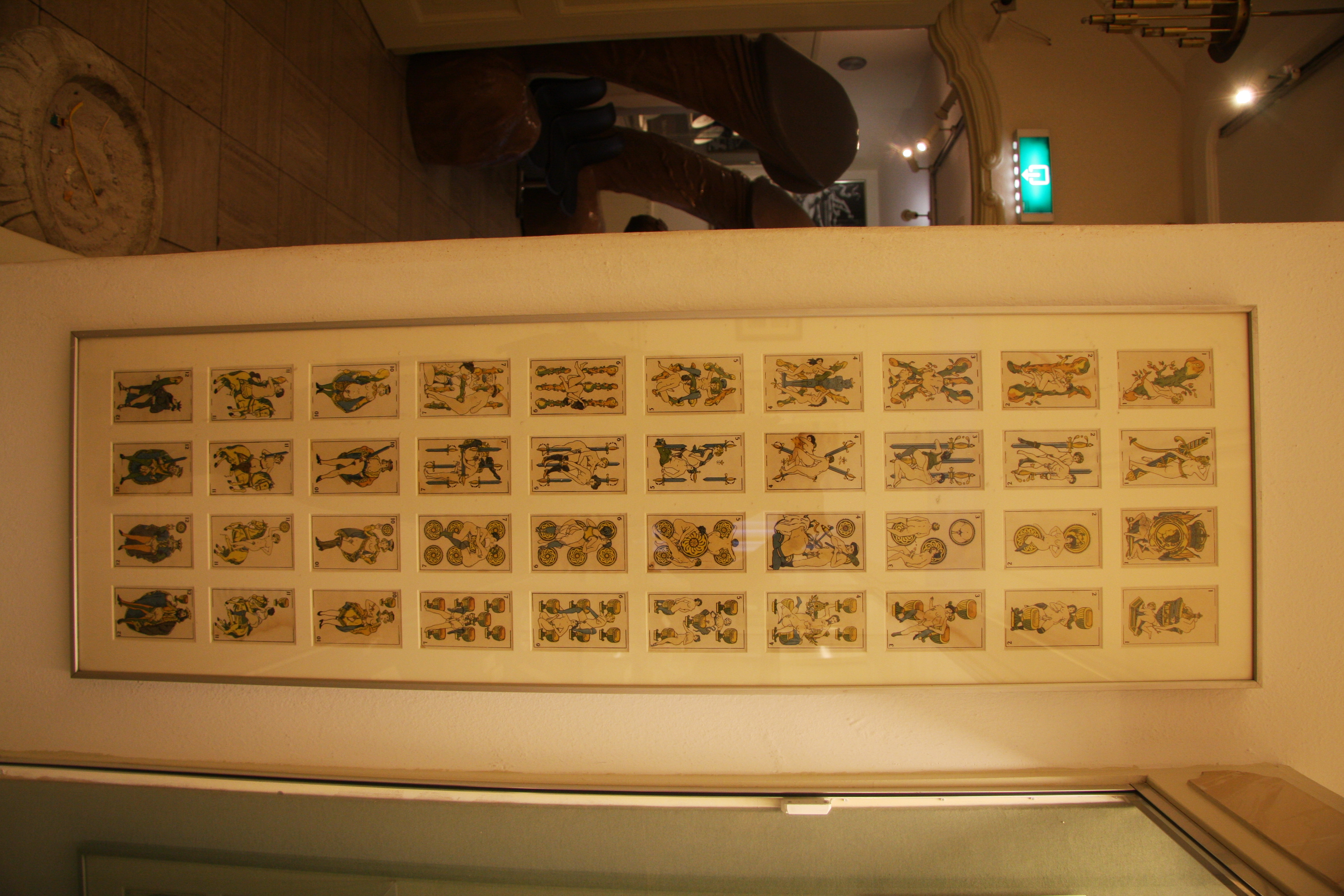 Framed grid of erotic illustrations on a wall inside Sex Museum Amsterdam, with dim lighting and additional exhibits visible in background