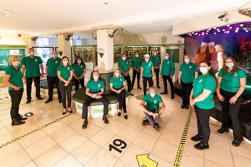Staff in green shirts pose in a museum interior with exhibition displays and numbered floor markers. The space features white tiled flooring and display cases.