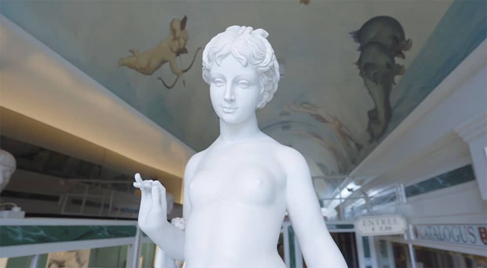 White statue of a woman in museum interior with decorative ceiling figures and glass display cases visible in the background