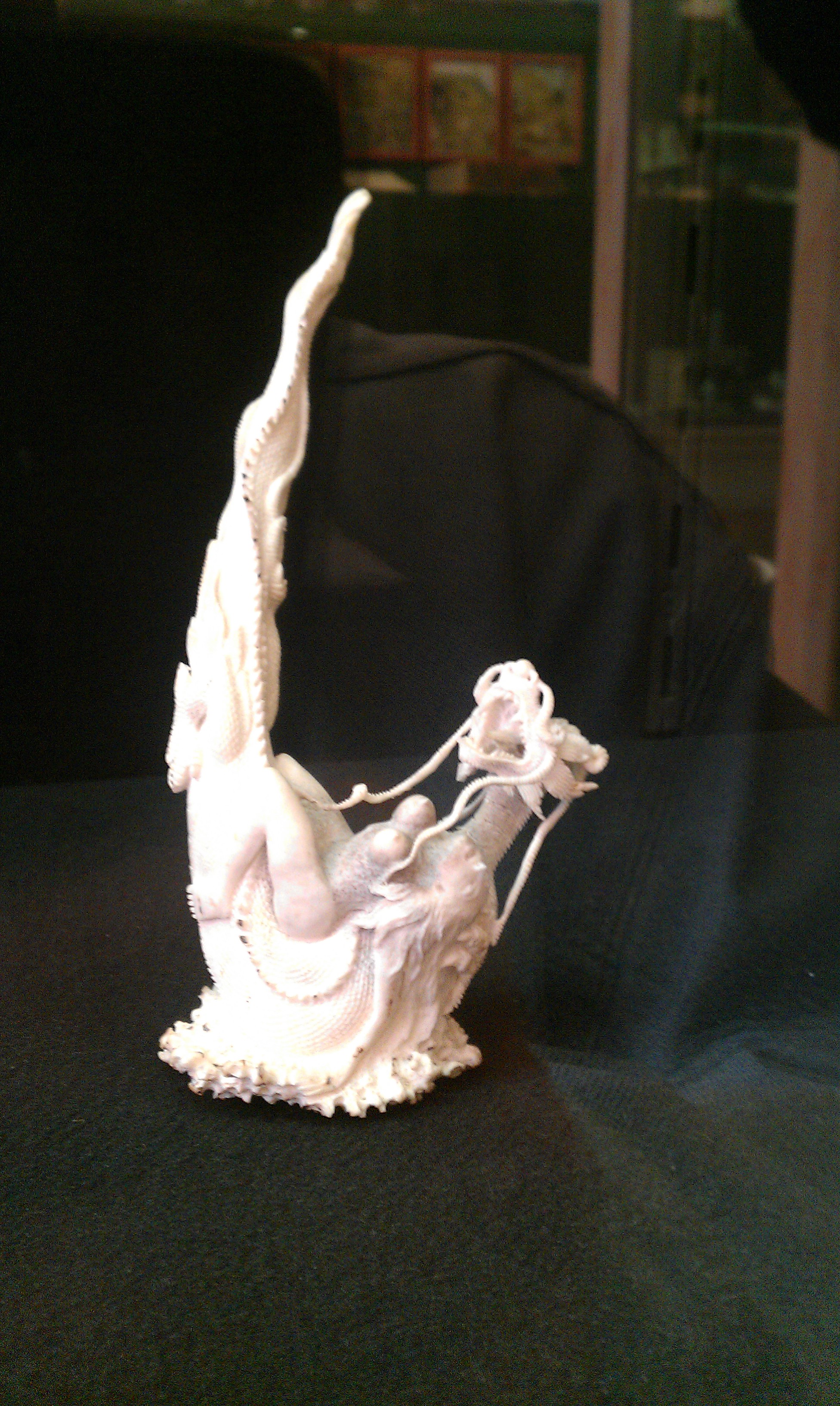 White detailed erotic sculpture with intertwined figures displayed on dark surface, likely museum exhibit