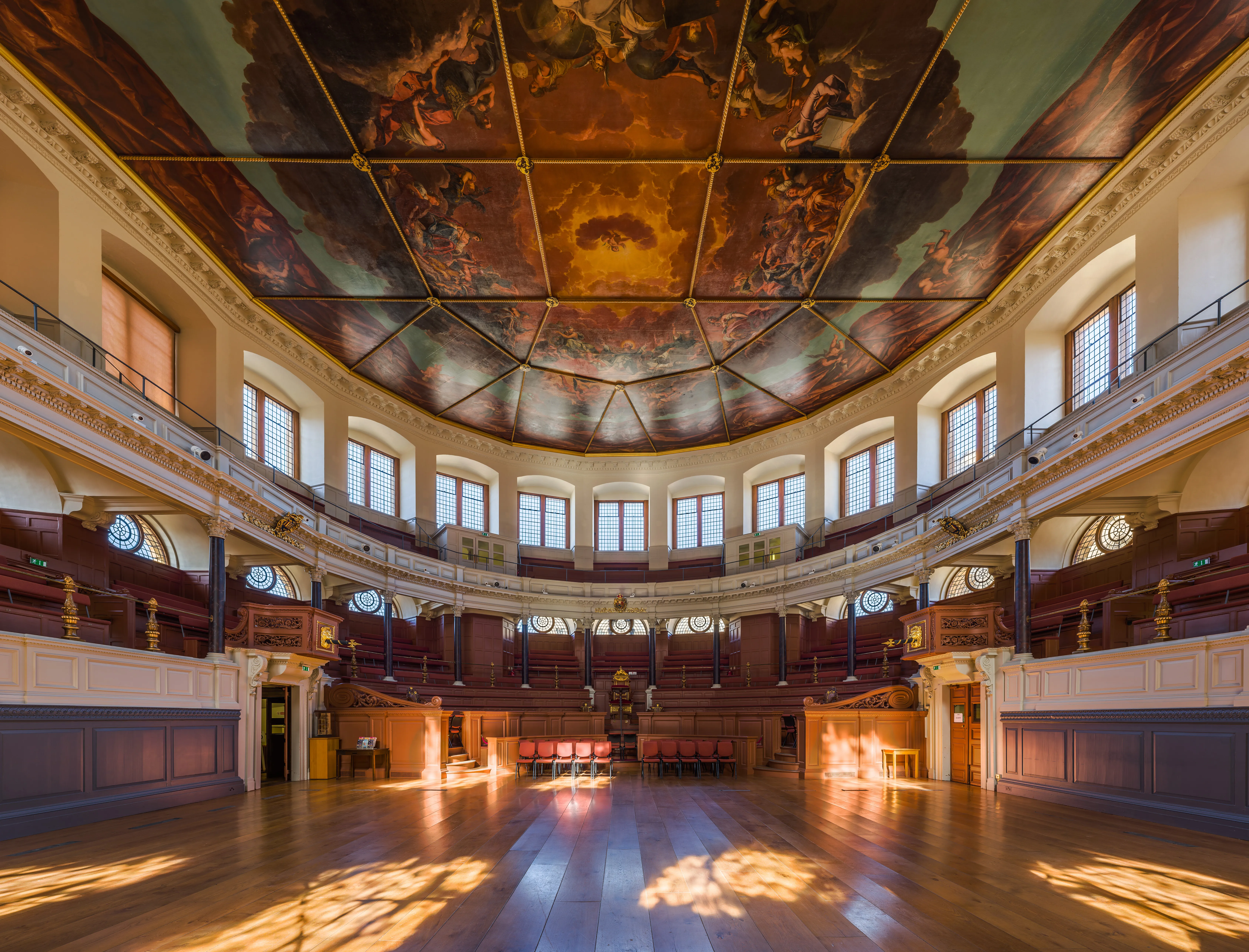 Interior del Sheldonian Theatre que muestra un vasto suelo de madera con luz solar entrando a través de ventanas arqueadas, galerías de madera escalonadas a lo largo de paredes curvas y un techo ornamentado y pintado con figuras clásicas.