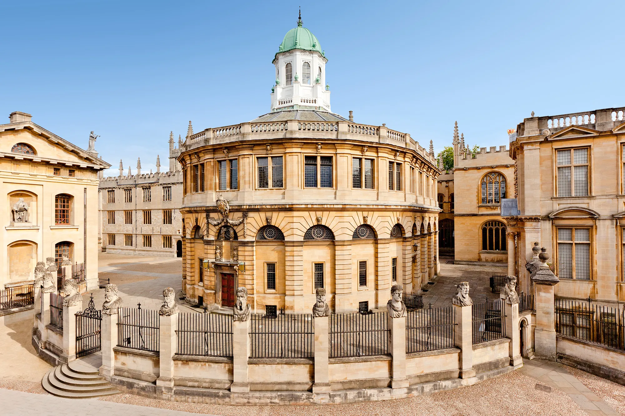 El Sheldonian Theatre, un edificio circular de piedra con una cúpula de techo verde, rodeado por una verja de hierro forjado con pilares de piedra y estatuas, en un día soleado.