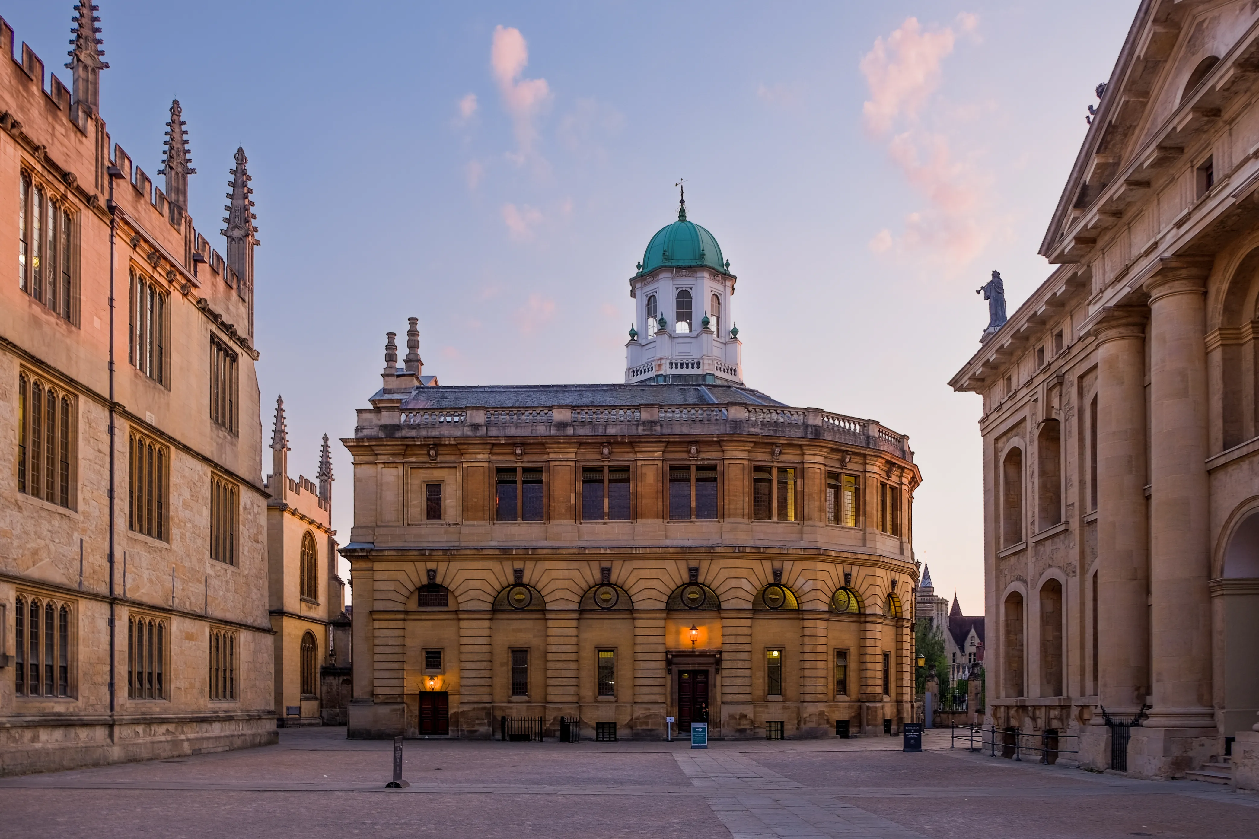 La fachada de piedra curvada del Sheldonian Theatre con su cúpula de cobre verde, flanqueada por edificios históricos de Oxford bajo un cielo crepuscular.