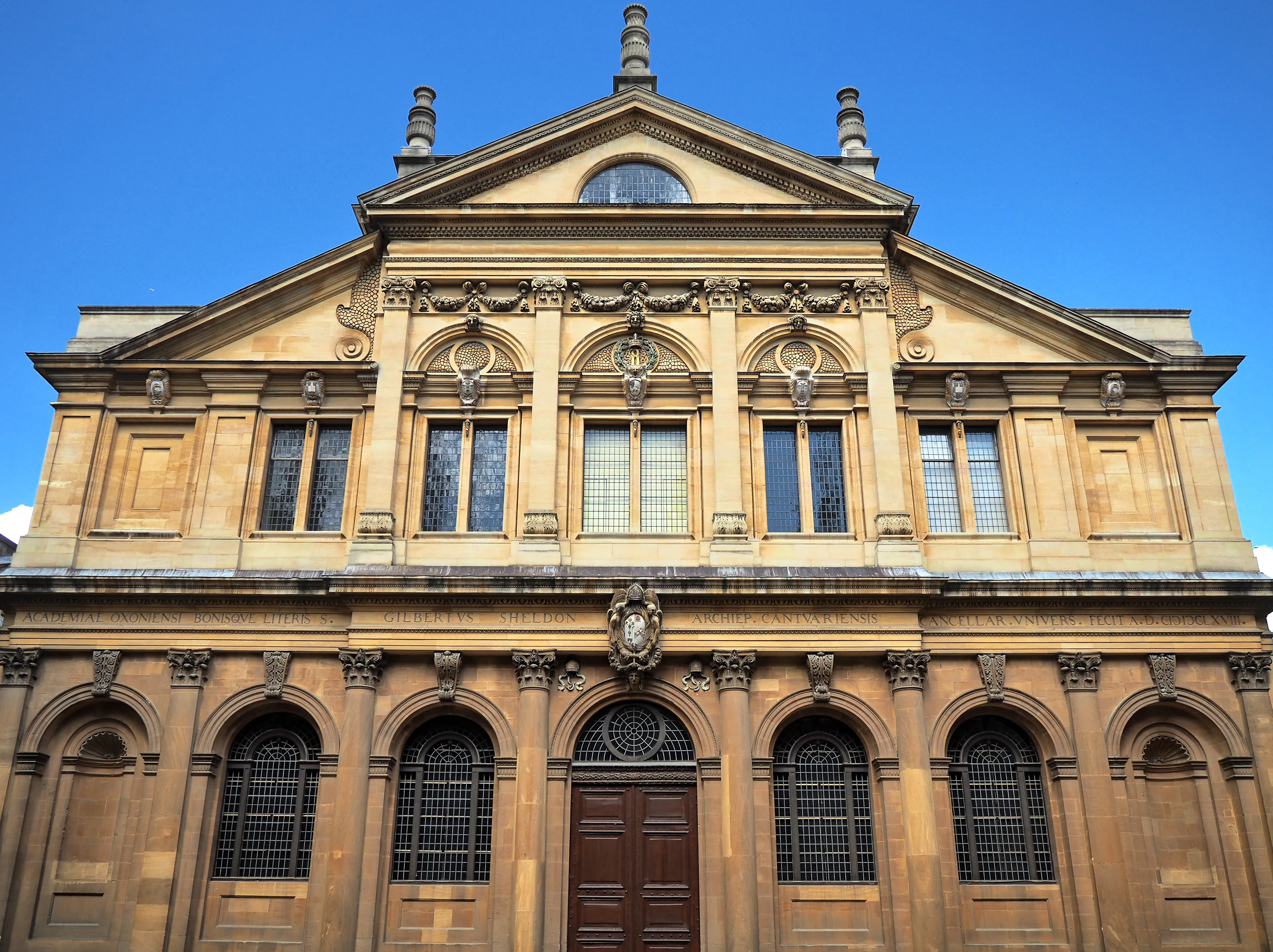 Fachada frontal del Sheldonian Theatre que muestra una arquitectura clásica de piedra con ventanas arqueadas, una puerta central de madera y un frontón triangular contra un cielo azul