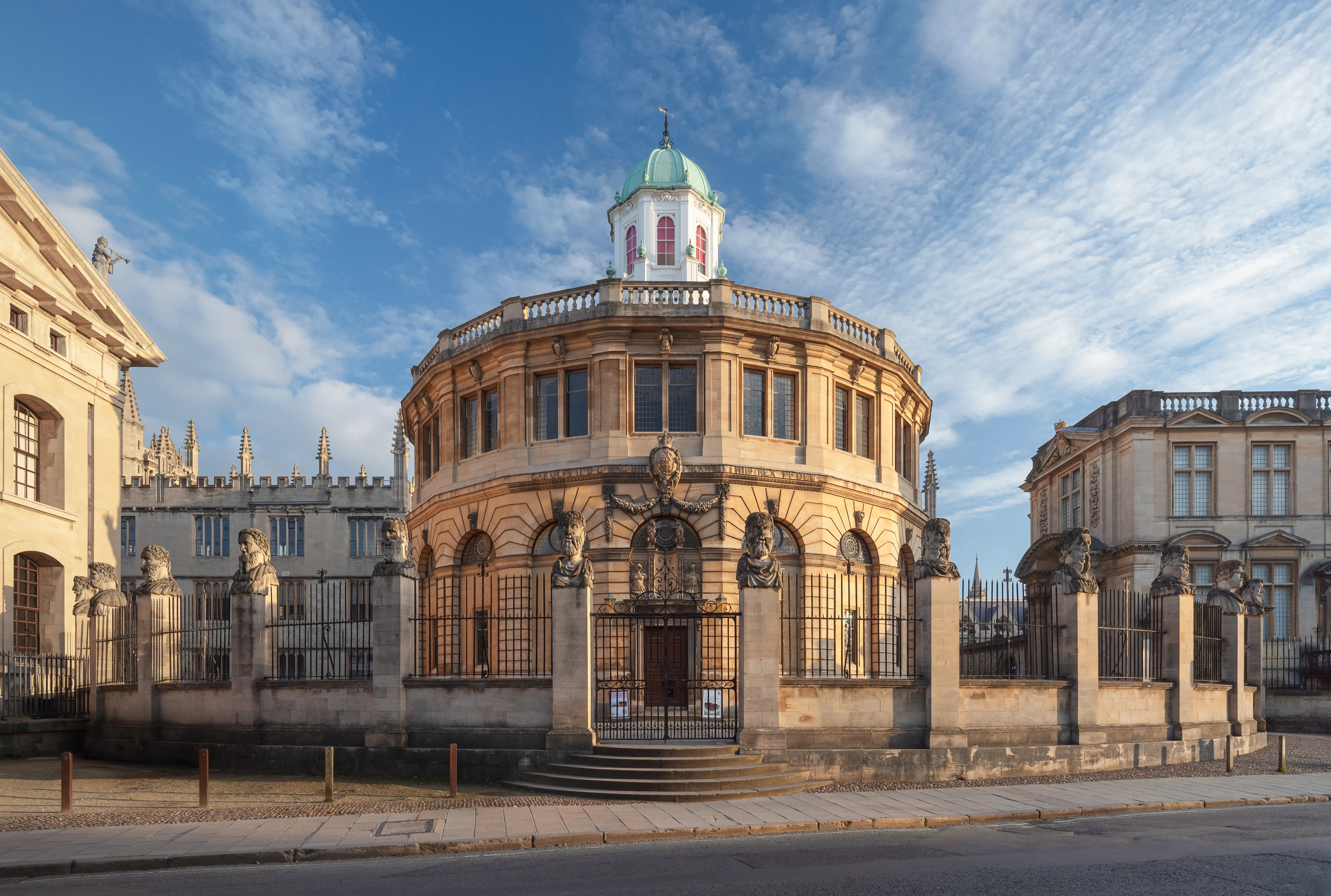 Vista exterior del Sheldonian Theatre en Oxford, que muestra su característica fachada de piedra ovalada con ventanas en arco, su entrada ornamentada y su cúpula de cobre verde bajo un cielo azul