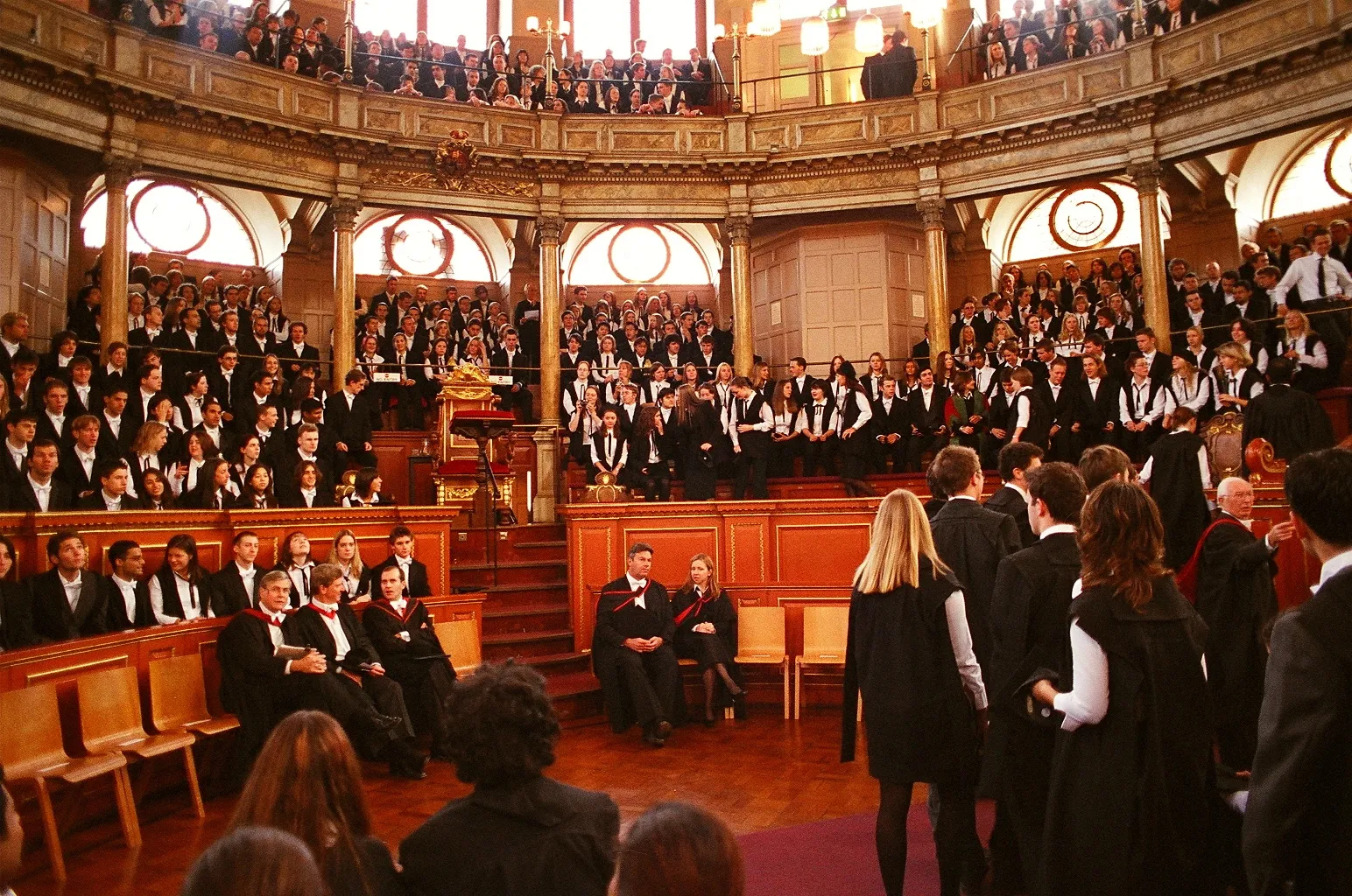 Interior del Sheldonian Theatre lleno de personas con togas académicas negras durante una ceremonia de matriculación, mostrando asientos de madera en niveles, ventanas arqueadas y paneles de madera ornamentados.