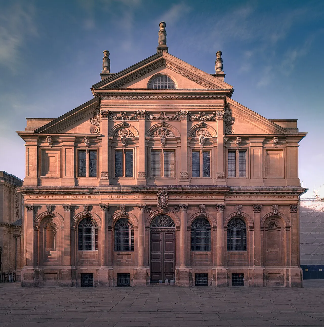 Fachada frontal del Sheldonian Theatre que muestra un edificio de piedra de estilo clásico con un frontón triangular, estatuas en la línea del tejado, ventanas arqueadas en la planta baja y una puerta de entrada central bajo un cielo azul.