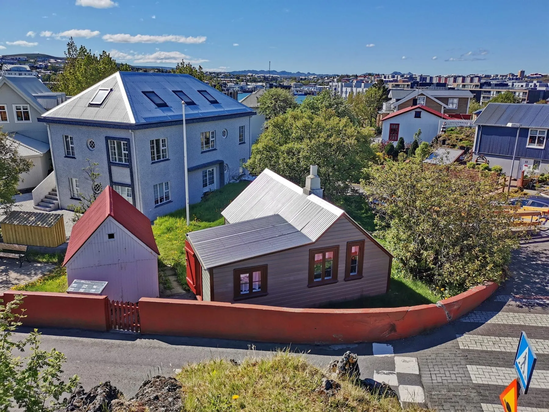 Luchtfoto van een klein historisch huis van golfplaat met een gebogen rood hek, omringd door moderne woongebouwen onder een helderblauwe lucht in Hafnarfjörður
