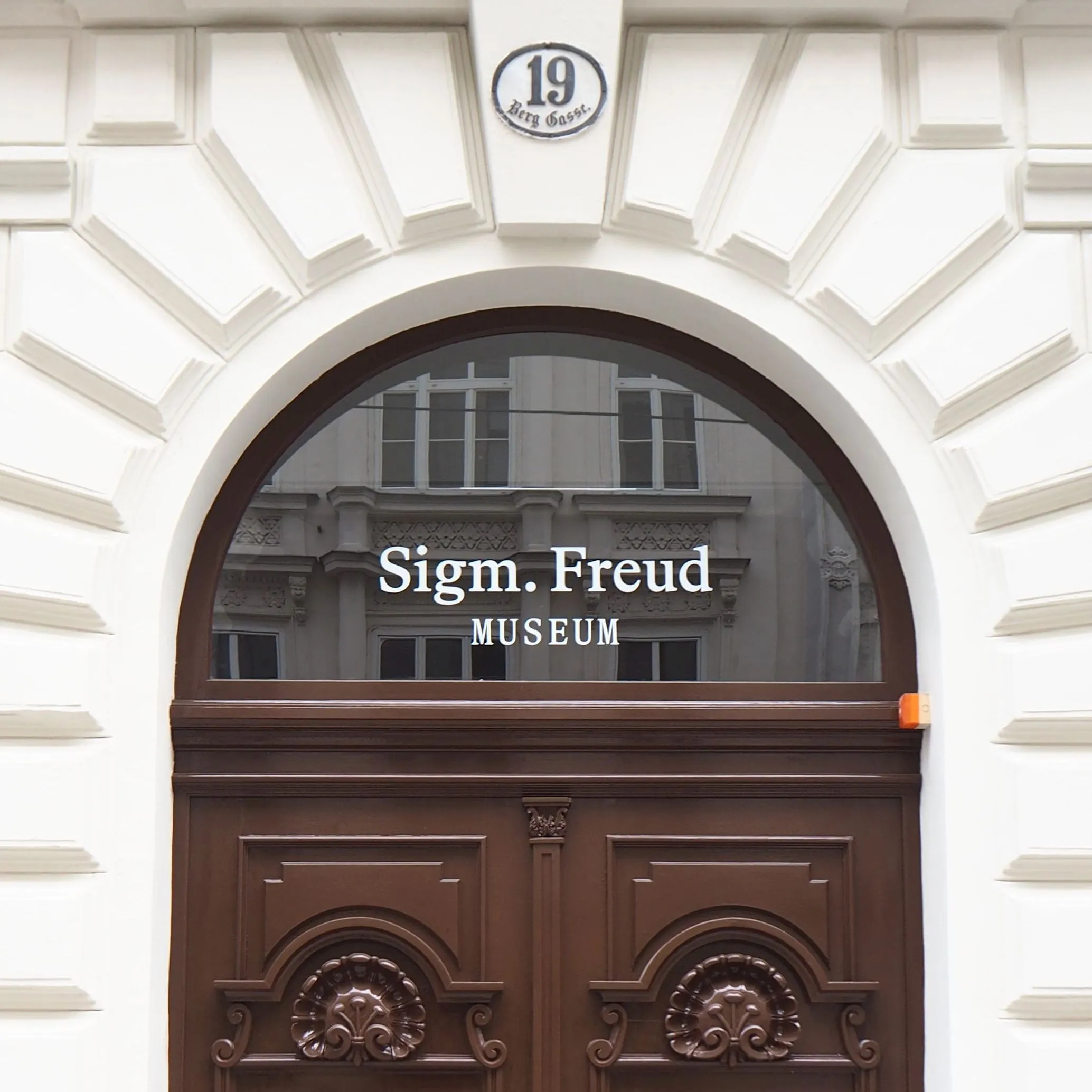 Bruine houten deur met gebogen glas-in-lood paneel met de tekst 'Sigm. Freud MUSEUM', geplaatst in een witte gevel met nummer 19-plaquette erboven