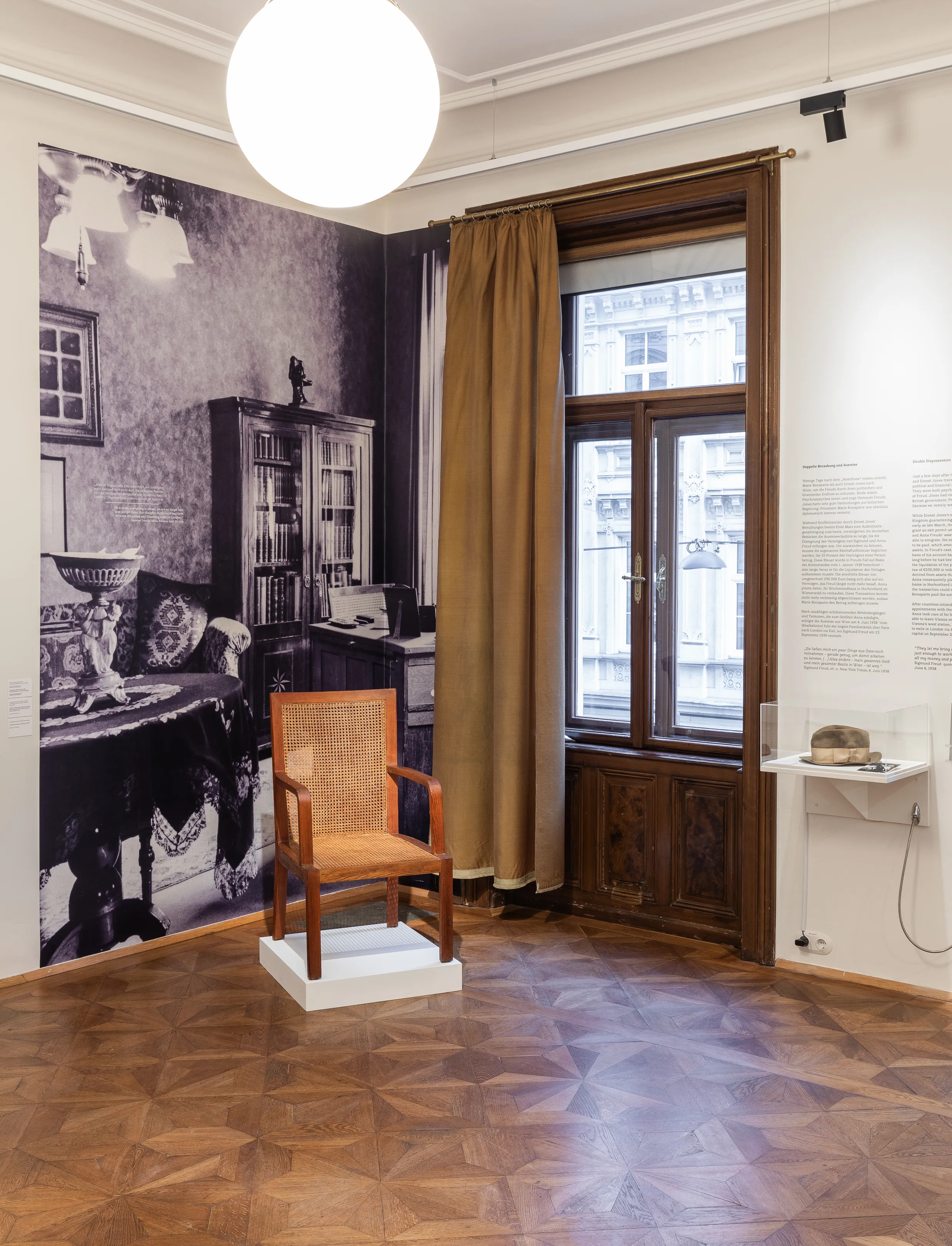 interieur van een museumzaal met een grote zwart-witfoto van een studeerkamer, een houten stoel op een wit voetstuk en een raam met bruine gordijnen