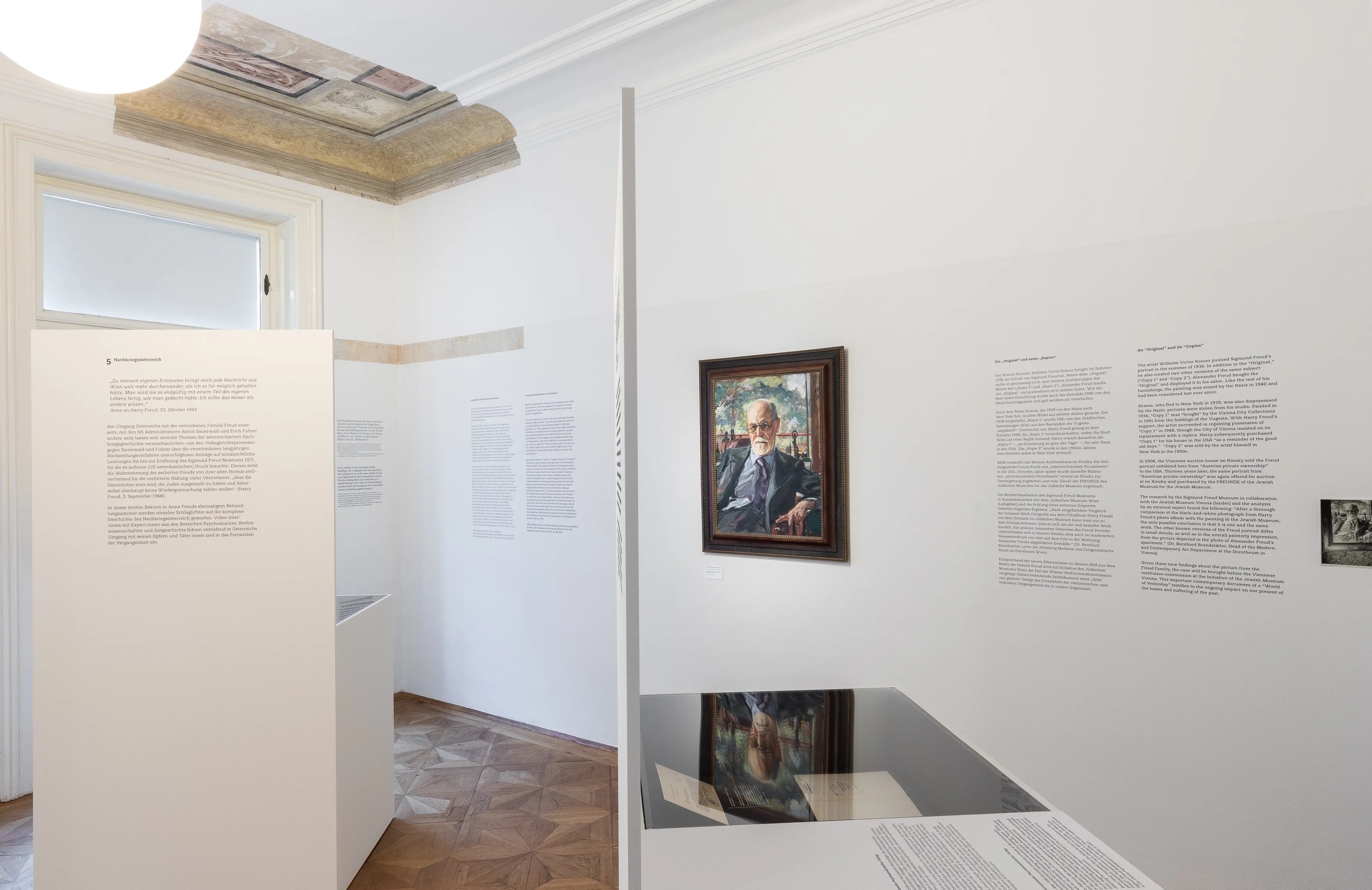 Een ingelijst portret van Sigmund Freud aan een witte muur, omgeven door informatieborden en een reflecterend oppervlak eronder.