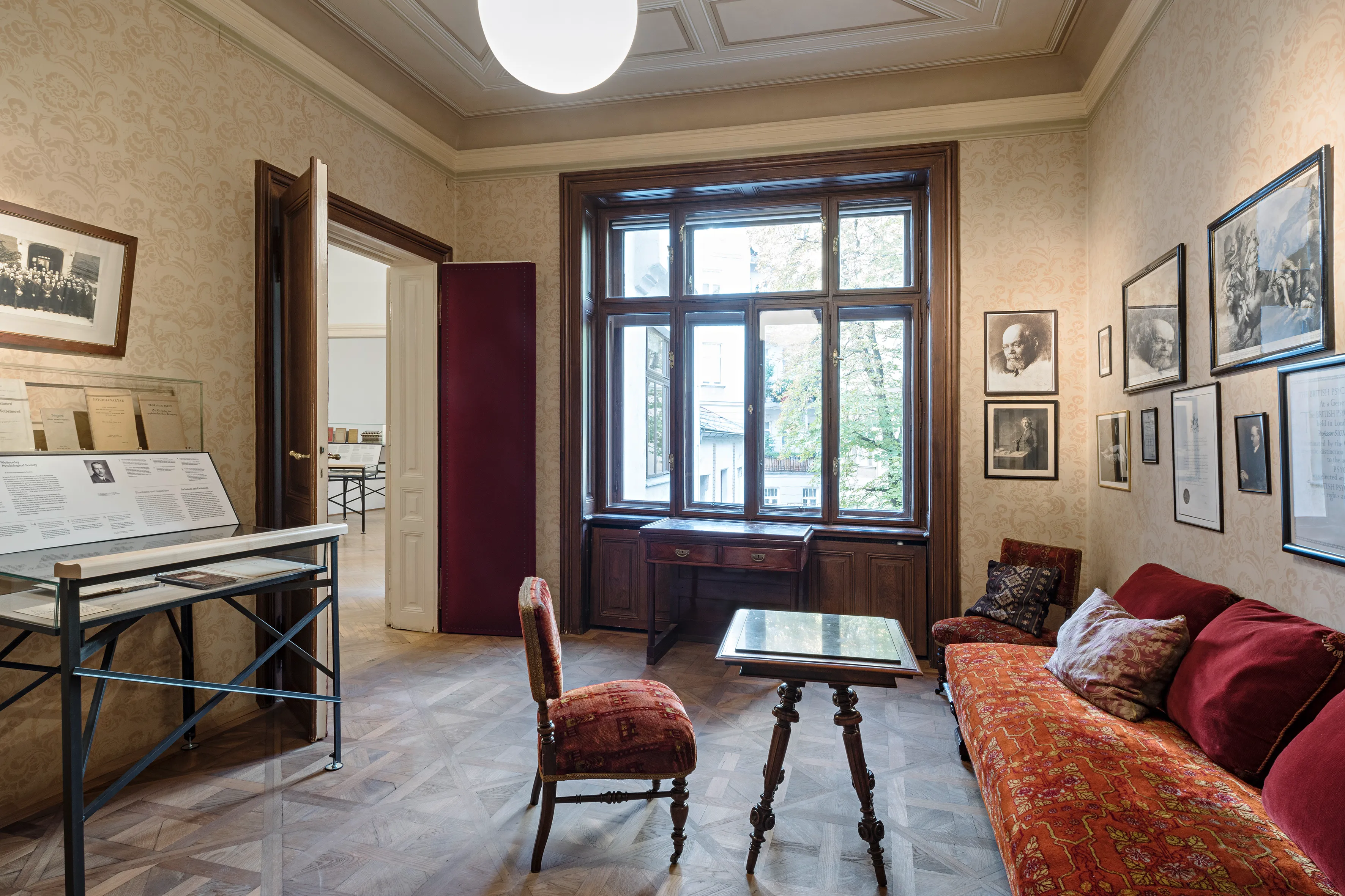 Interieur van het Sigmund Freud Museum met een rode bank met patroon, een kleine tafel, een stoel en ingelijste foto's aan de muren