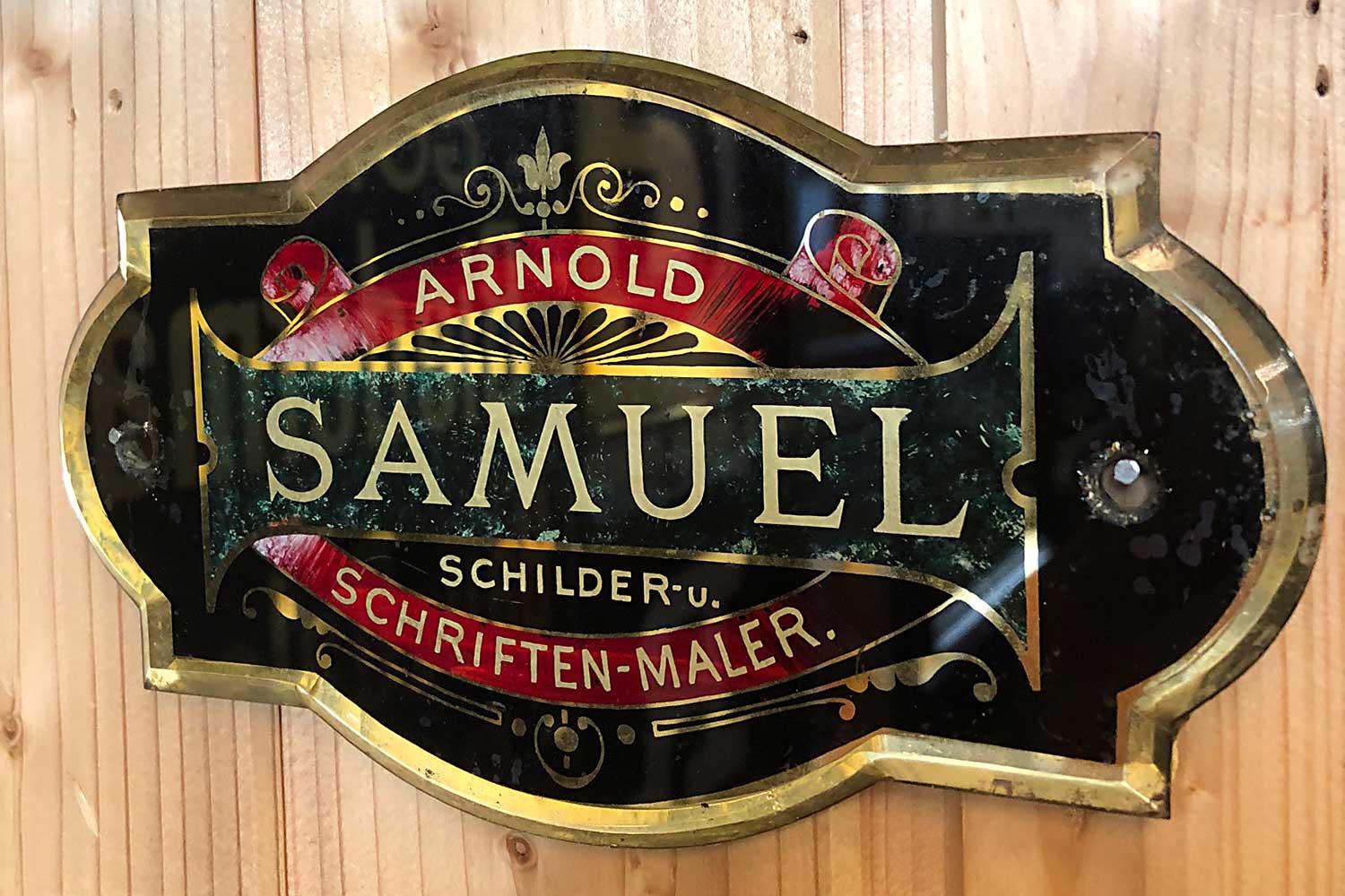 Un letrero decorativo con letras doradas sobre un fondo negro, que dice 'ARNOLD SAMUEL SCHILDER-u. SCHRIFTEN-MALER' con adornos ornamentales y acentos rojos.