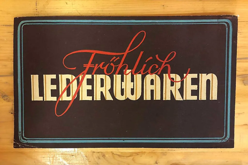 Een handgeschilderd bord met de tekst 'Fröhlich LEDERWAREN' in rood cursief en beige blokletters op een donkere achtergrond