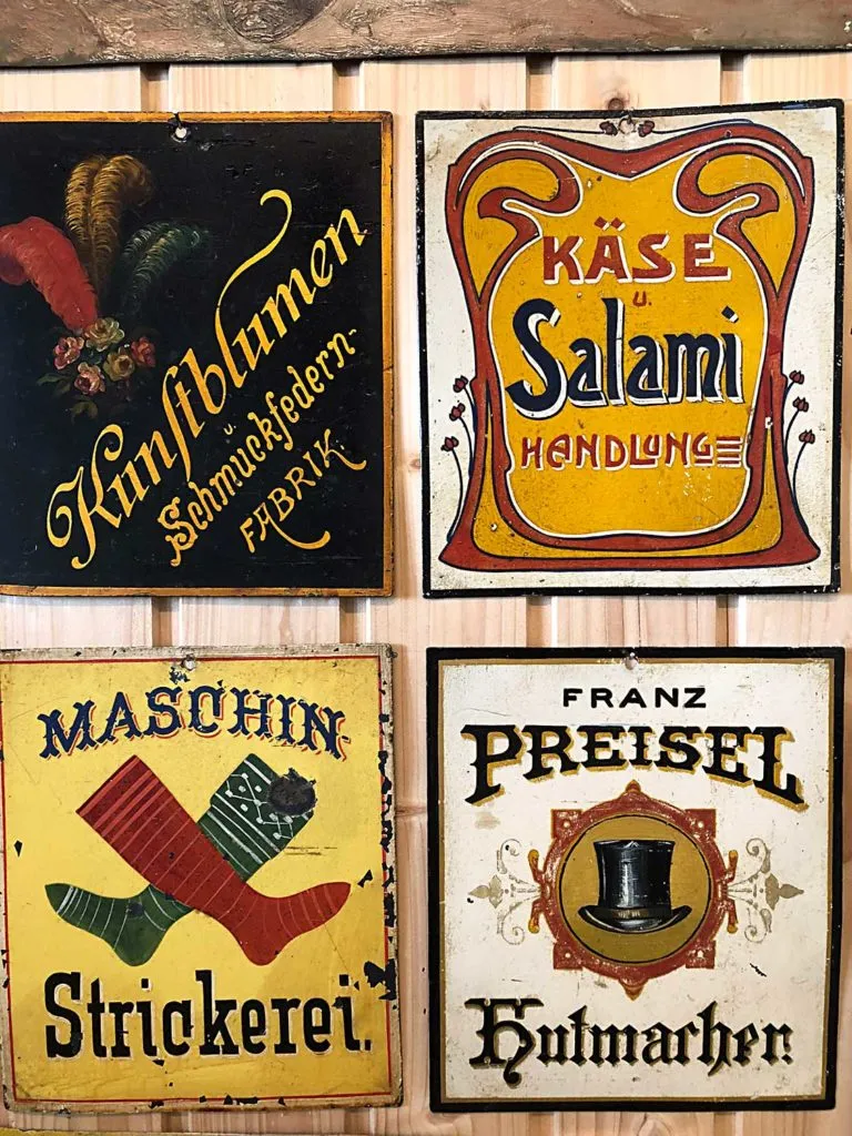 Cuatro letreros vintage pintados a mano con letras y ilustraciones ornamentadas, que incluyen 'Kunstblumen', 'Käse Salami', 'Maschin Strickerei' y 'Franz Preisel Hutmarken', montados en una pared de madera.