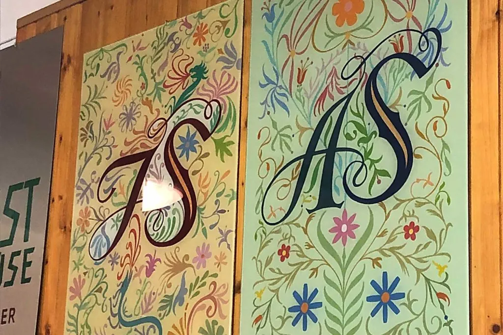 Dos letreros coloridos pintados a mano con letras elaboradas y diseños florales en una pared de madera