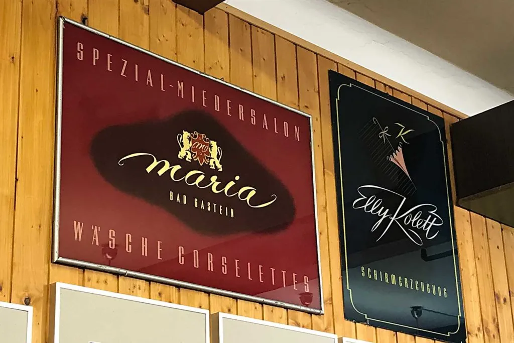 Dos letreros pintados a mano sobre una pared de madera, uno rojo con letras doradas que dicen 'maria' y otro negro con letras blancas y escritura a mano que dice 'Elly Kost'.