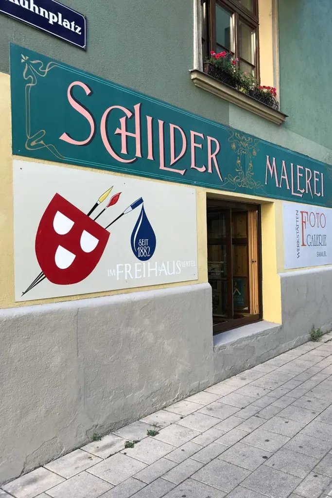Pared exterior de un edificio con un gran letrero que dice 'Schilder Malerei' en letras rosas sobre un fondo verde, una máscara de teatro roja con pinceles y una señal de calle azul encima de la entrada