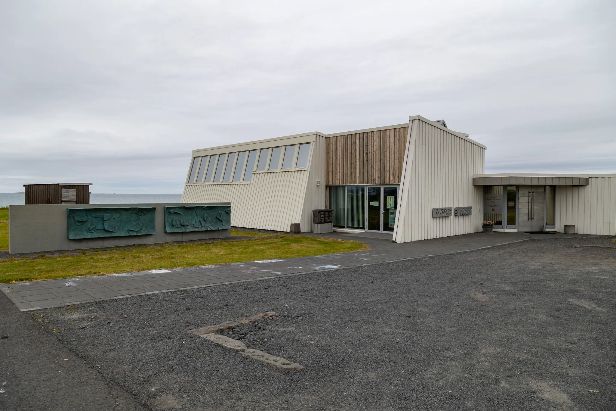 Vista exterior del Sigurjón Ólafsson Museum, un edificio blanco modernista con arquitectura angular, revestimiento de madera y esculturas en relieve verdes sobre un muro bajo, con el océano visible al fondo.