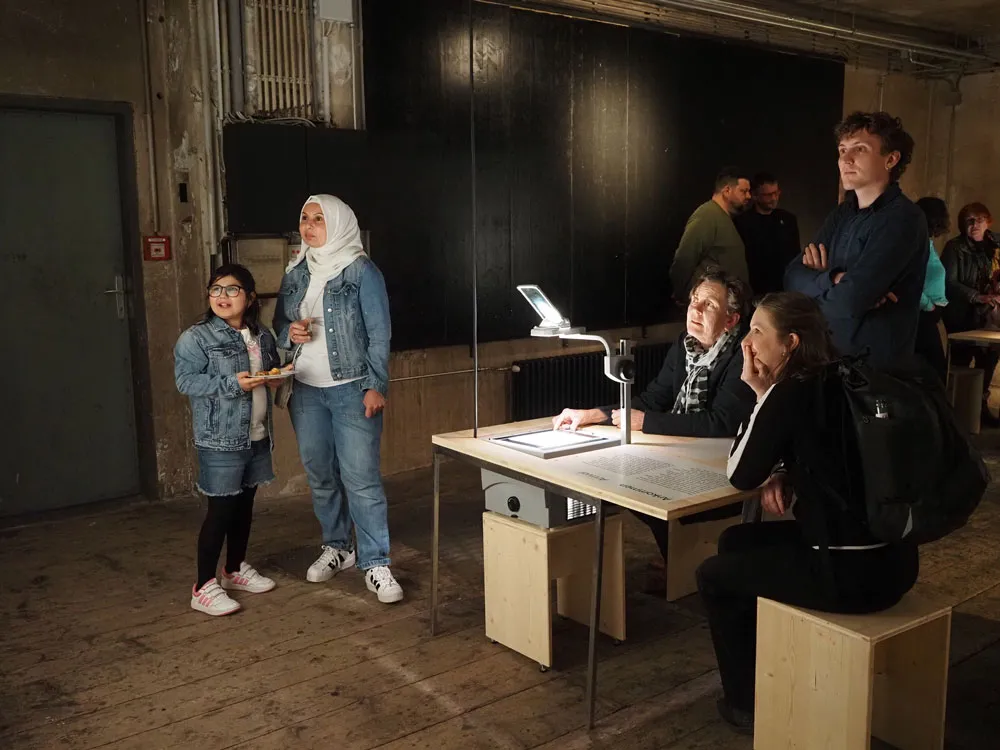 People viewing an interactive exhibit with a digital display on a table in a gallery with wooden floors and dark walls