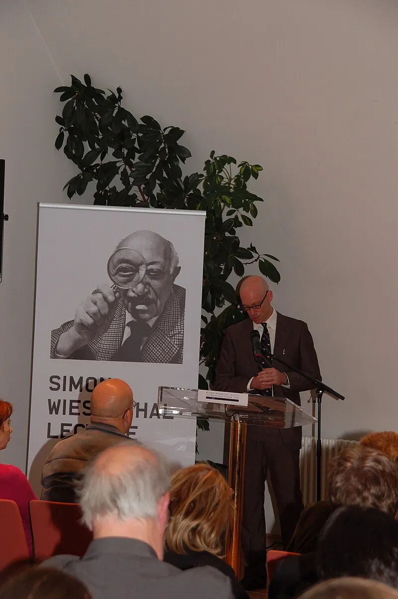 A man speaking at a podium in front of a banner with a portrait of Simon Wiesenthal and the text 'SIMON WIESENTHAL LECTURE' in a room with an audience.