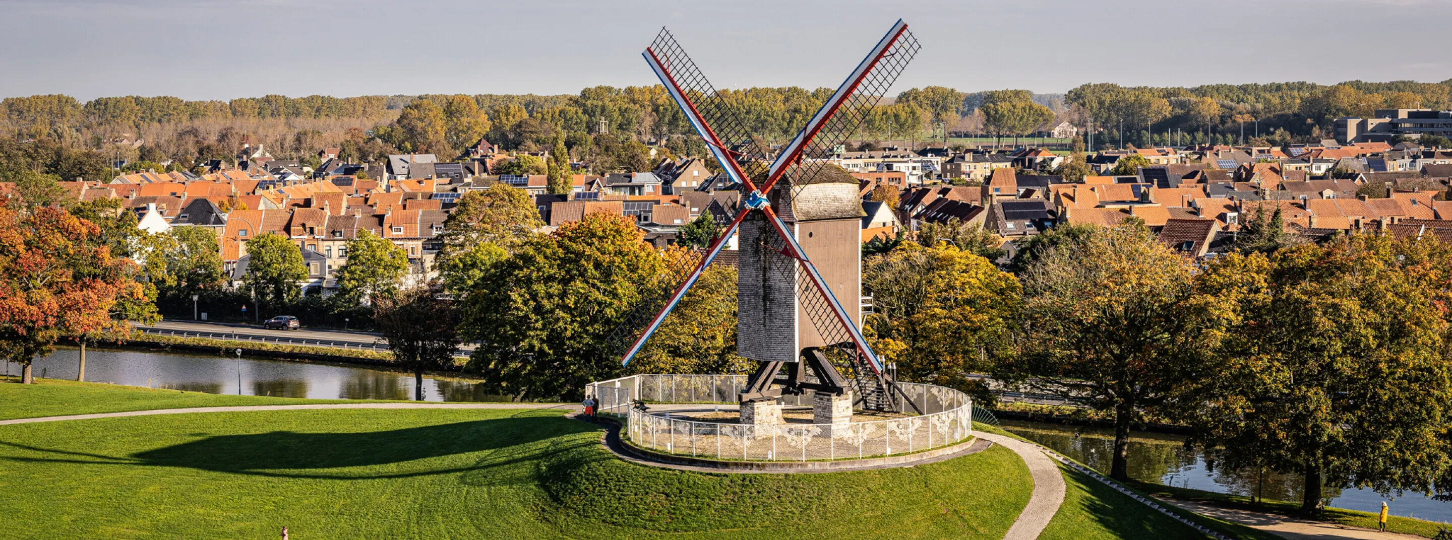 Panoramiczny widok na wiatrak Sint-Janshuismolen z biało-czerwonymi skrzydłami na trawiastym wzgórzu, z widokiem na panoramę Brugii, jesienne drzewa i kanał.