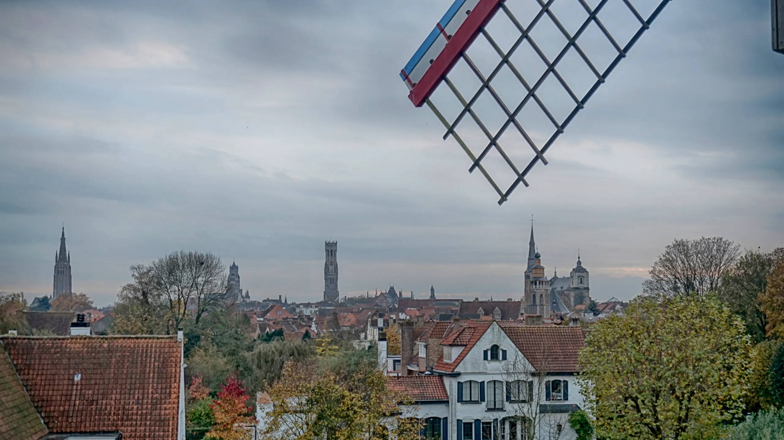 Widok z wiatraka Sint-Janshuismolen na panoramę Brugii z wieżami kościelnymi, dzwonnicą Belfort, dachami z czerwoną dachówką i jesiennymi drzewami, z widocznym w prawym górnym rogu skrzydłem wiatraka.