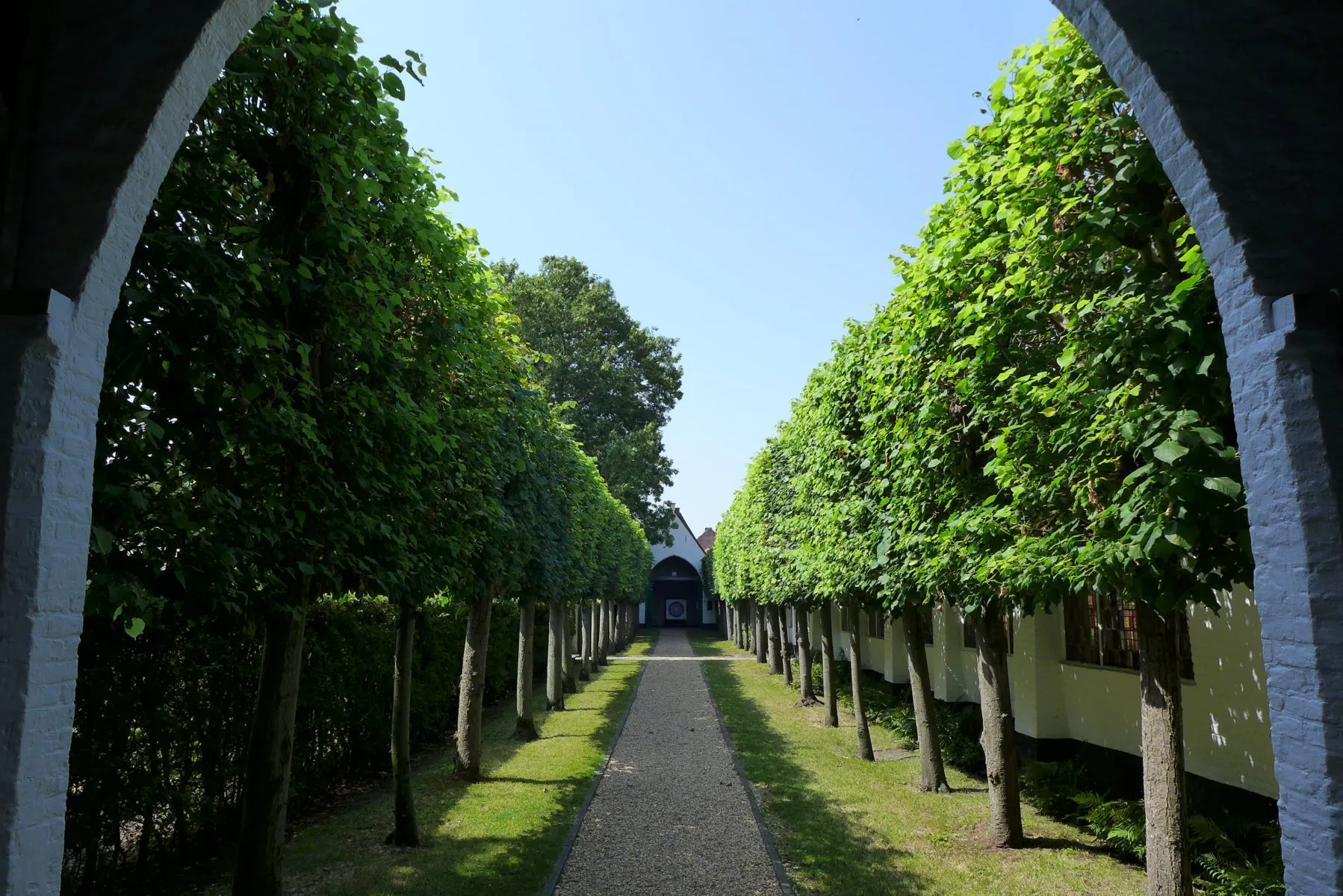 Un sendero de grava bordeado de árboles y enmarcado por un arco blanco conduce hacia un edificio al final del jardín de tiro con arco del gremio, con árboles cuidadosamente recortados a ambos lados bajo un cielo azul despejado.