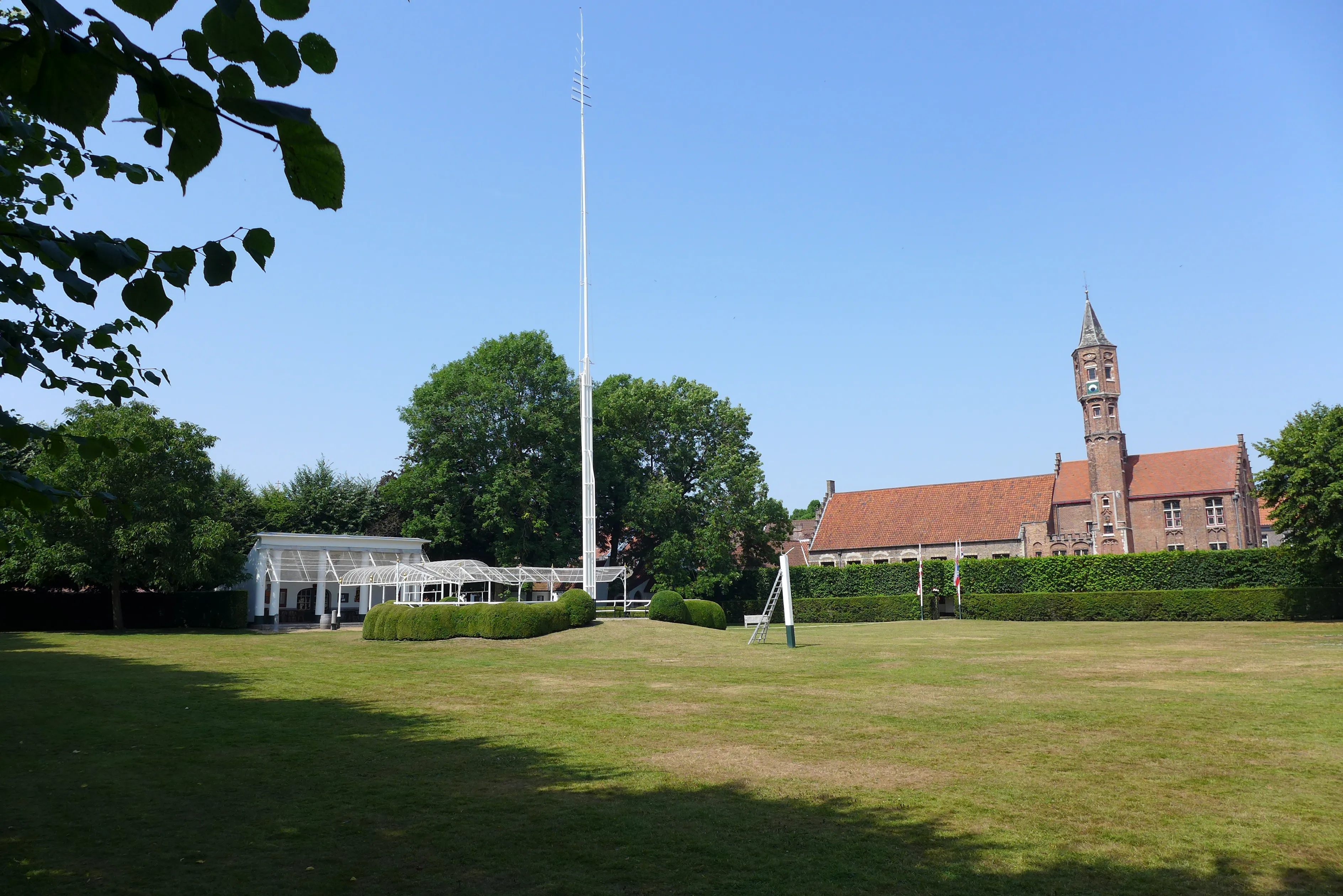 Grasrijke boogschutterstuin met een hoge witte vlaggenmast, een witte paviljoentent aan de linkerkant en een roodstenen gildehuis met een klokkentoren aan de rechterkant onder een helderblauwe lucht