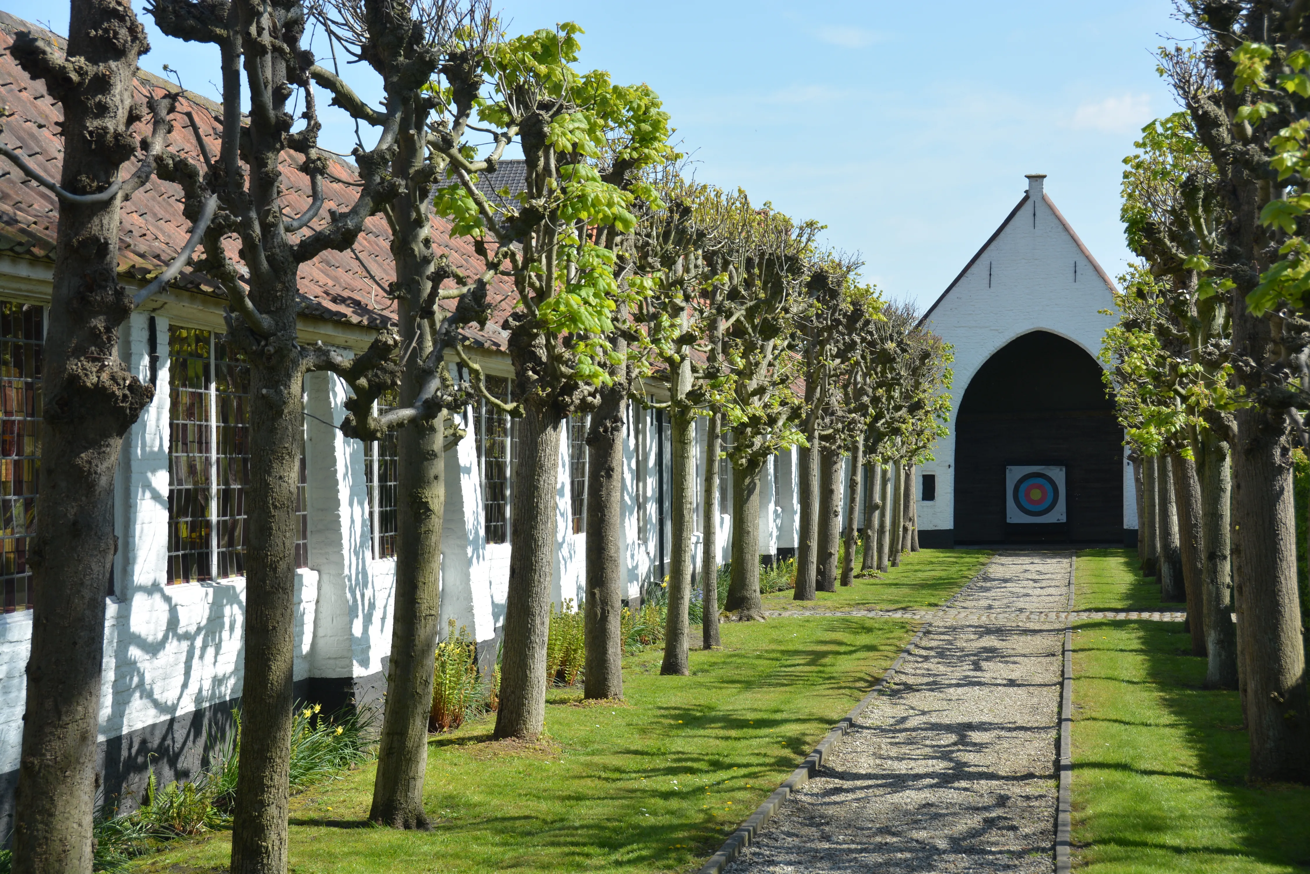 Een klinkerpad met gekandelaberde bomen leidt naar een wit gebouw met een grote boogingang waar een kleurrijk schietdoel hangt.