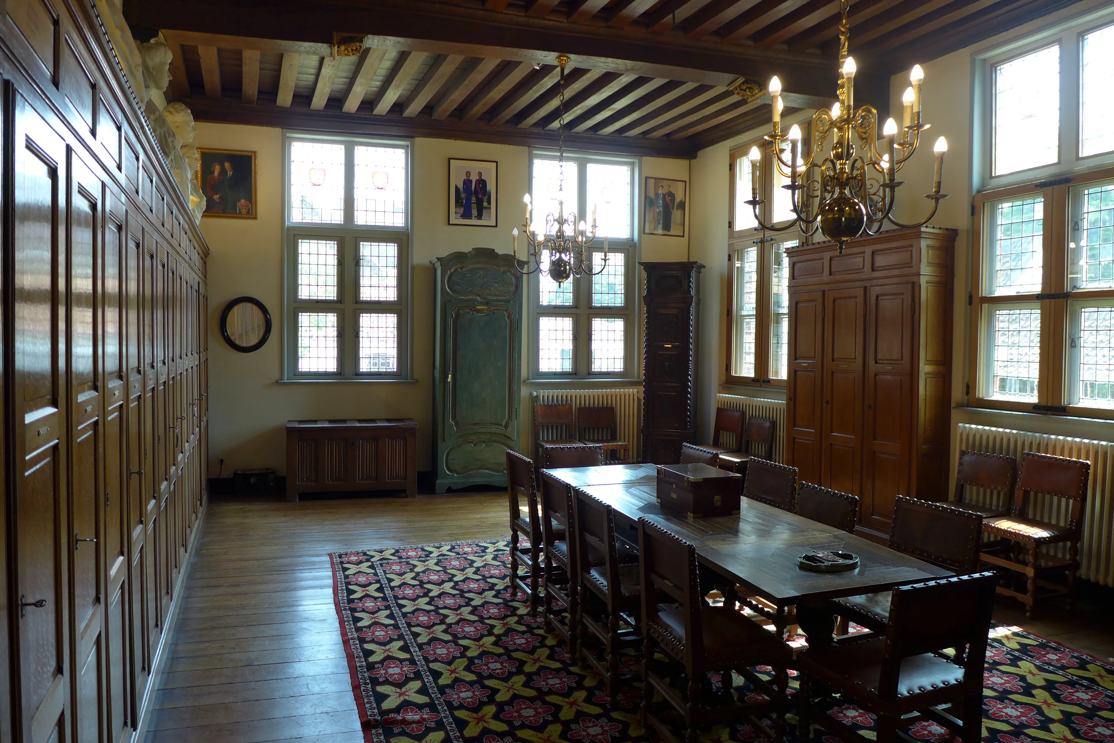 Interior de una histórica sala de gremio que cuenta con una larga mesa de madera con sillas, paneles de madera, una lámpara de araña y una alfombra estampada.