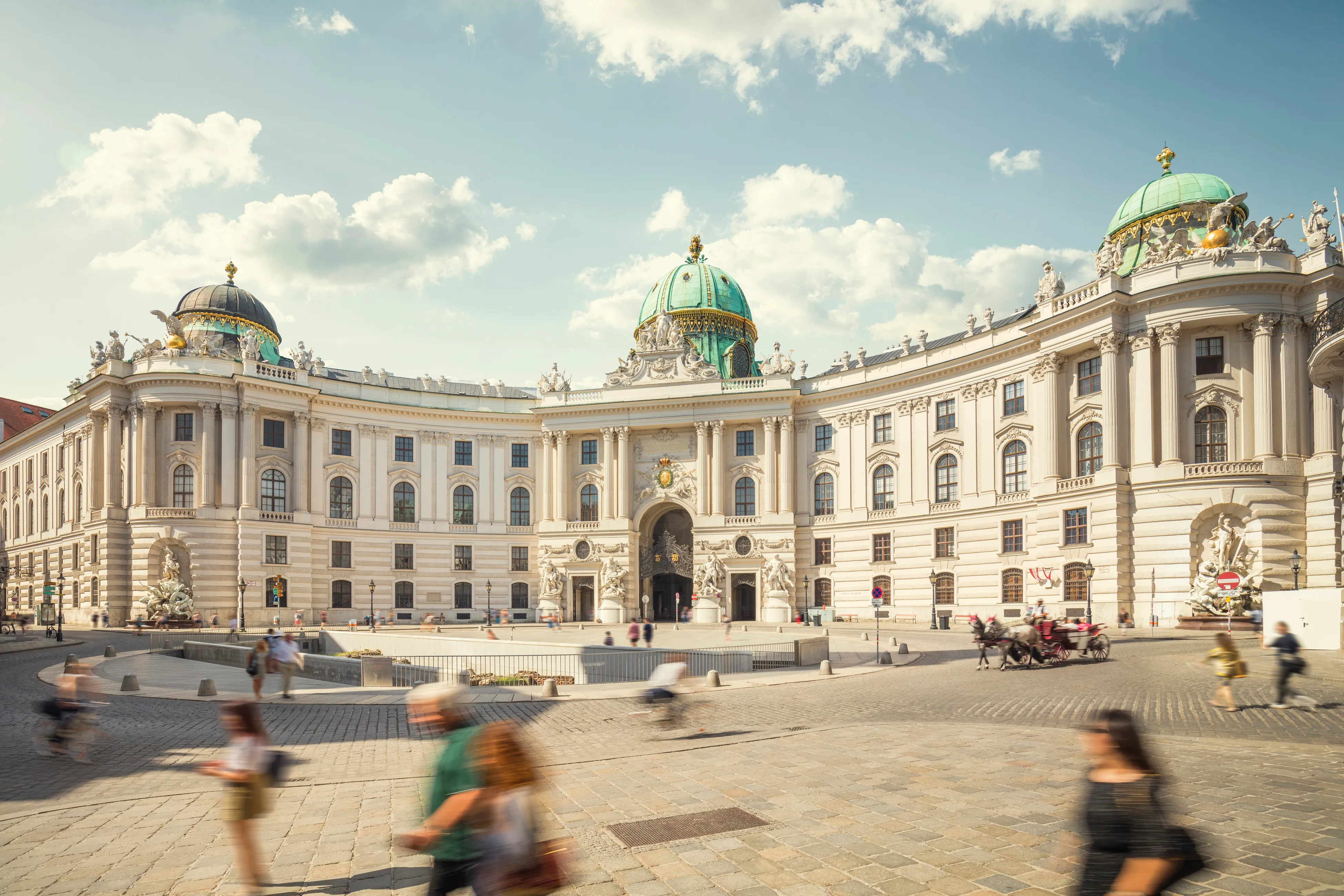 Muzeum Sisi, wspaniały barokowy pałac z fasadą z białego kamienia, zielonymi kopułami i licznymi oknami, z ludźmi spacerującymi po placu na pierwszym planie.