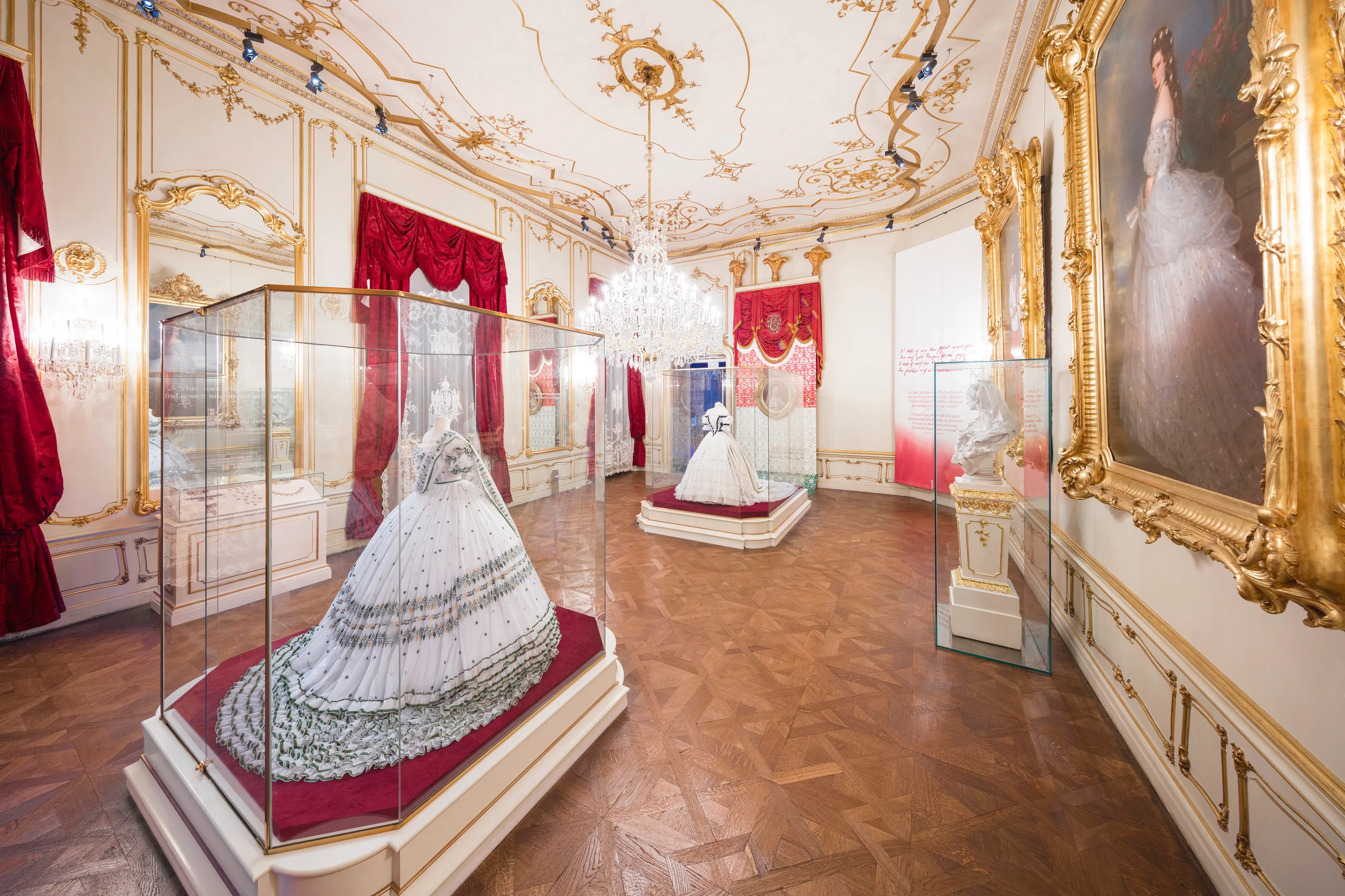 Een museumgalerij met een witte japon in een glazen vitrine, een groot portret van een vrouw in een vergulde lijst, en sierlijke goudomrande muren en plafond