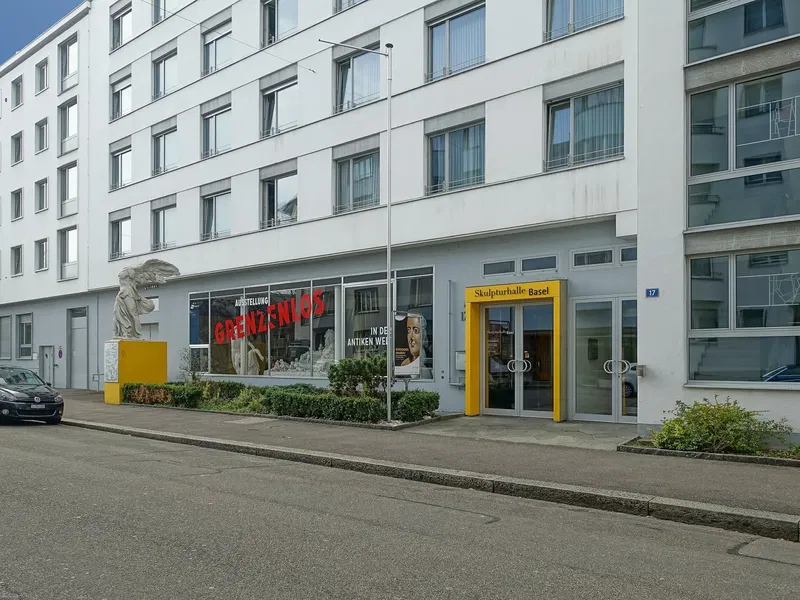Biały wielopiętrowy budynek z wejściem w żółtej ramie z napisem Skulpturhalle Basel, gipsowa rzeźba na żółtym cokole oraz plakaty wystawowe w oknach na parterze