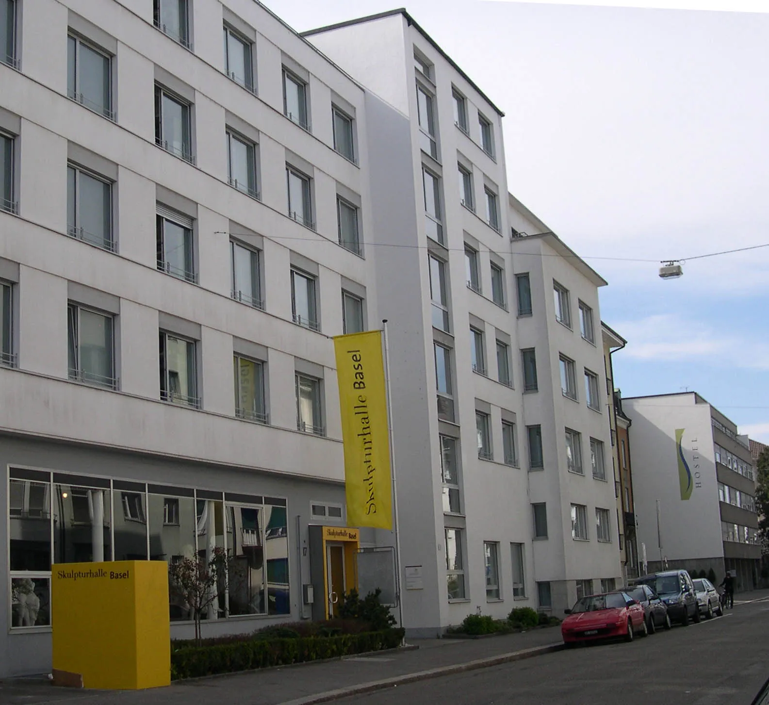 Widok z ulicy na biały, wielopiętrowy budynek Skulpturhalle Basel z żółtym pionowym banerem i żółtym oznakowaniem wejścia