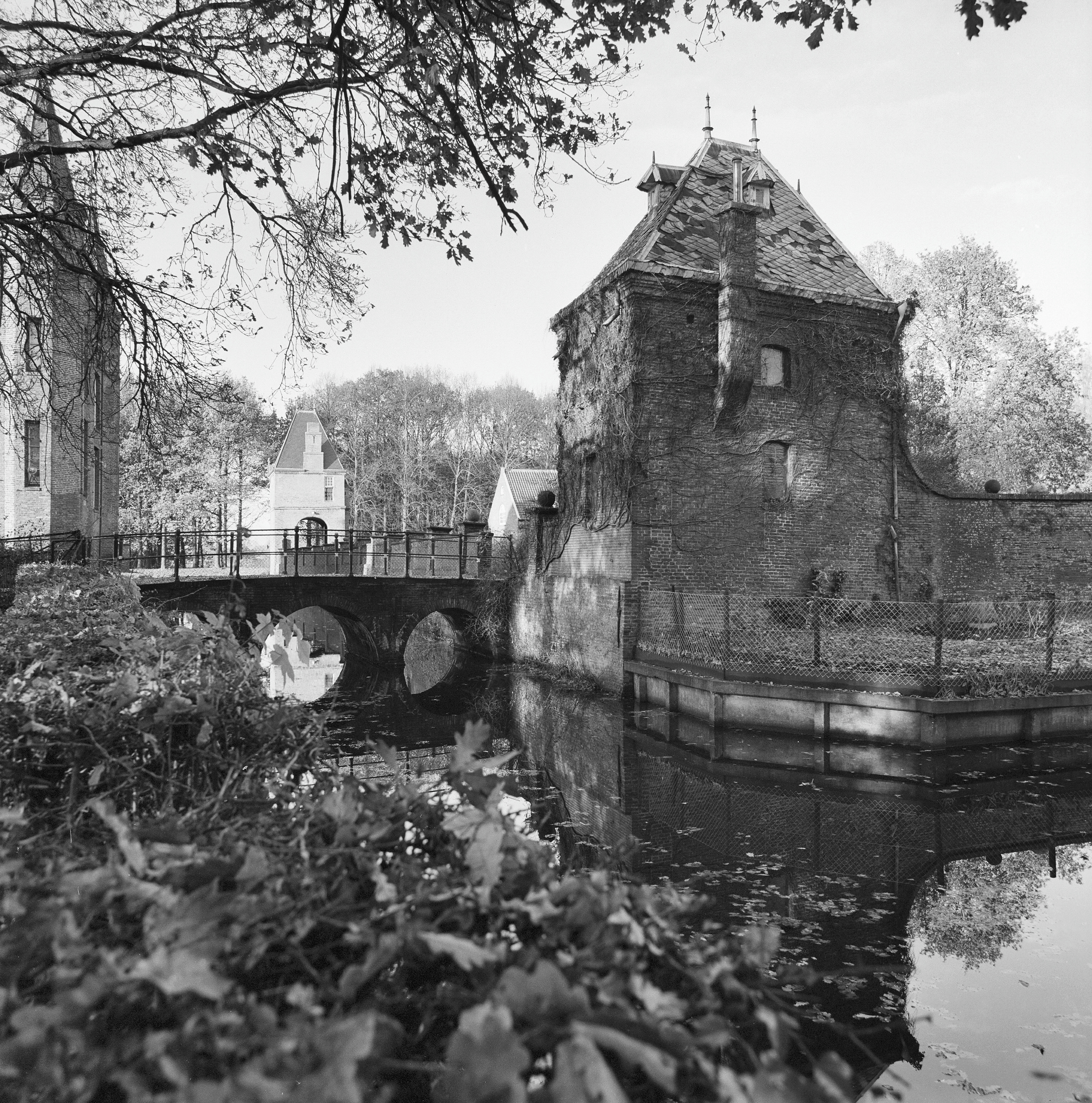 Black and white photograph of a historic stone castle tower with moat, arched bridge, and water reflection, surrounded by trees and foliage