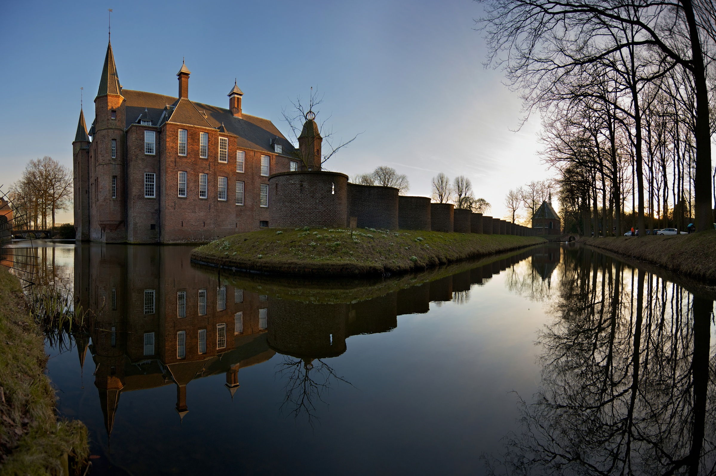 Red-brick historic castle with turrets and reflection in moat water, surrounded by trees and walls at dusk
