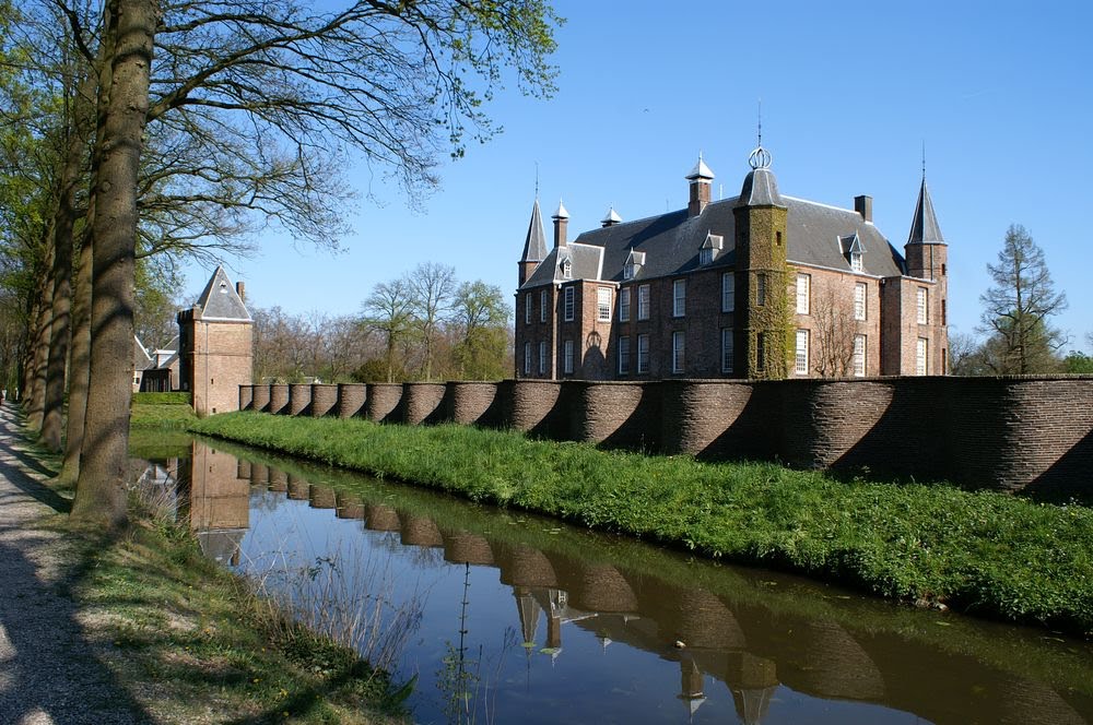 Historic brick castle with turrets and a moat, surrounded by greenery and a tree-lined path under a clear blue sky