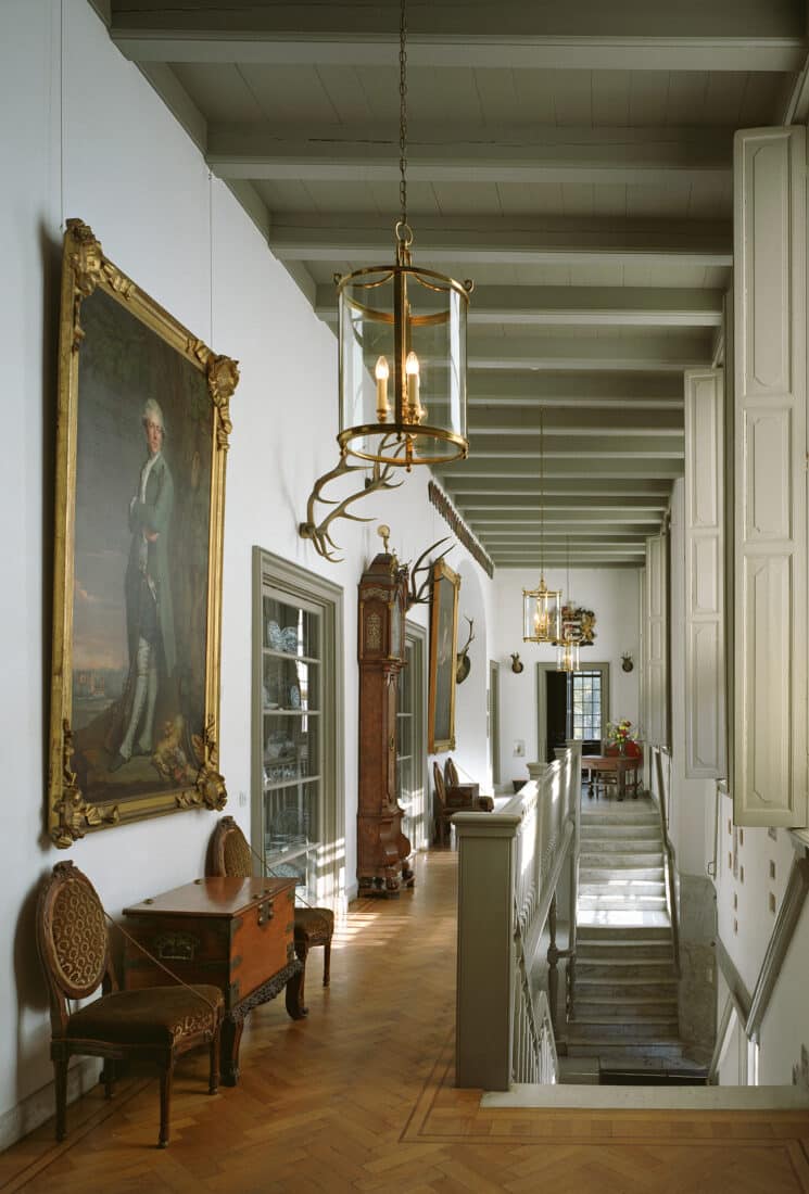 Long hallway in historic castle with wooden floors, period furniture, large paintings, and a staircase leading to another level