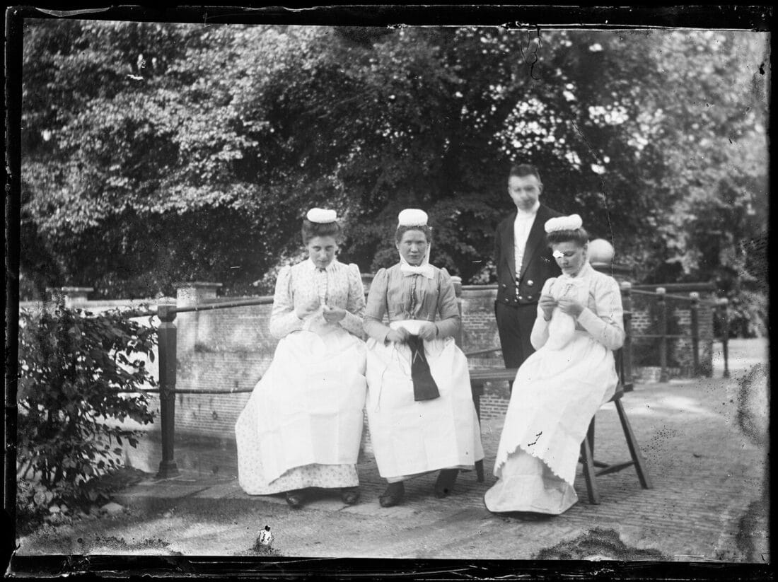 Three women in historical white dresses and caps sitting with a man standing behind them in an outdoor setting with trees