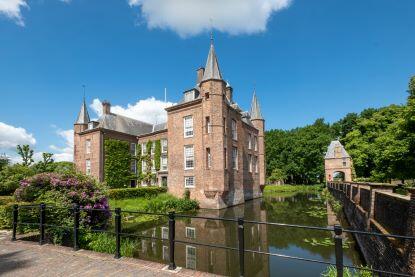 Red brick castle with turrets surrounded by a water moat under a bright blue sky with white clouds, featuring a bridge entrance and lush green landscaping
