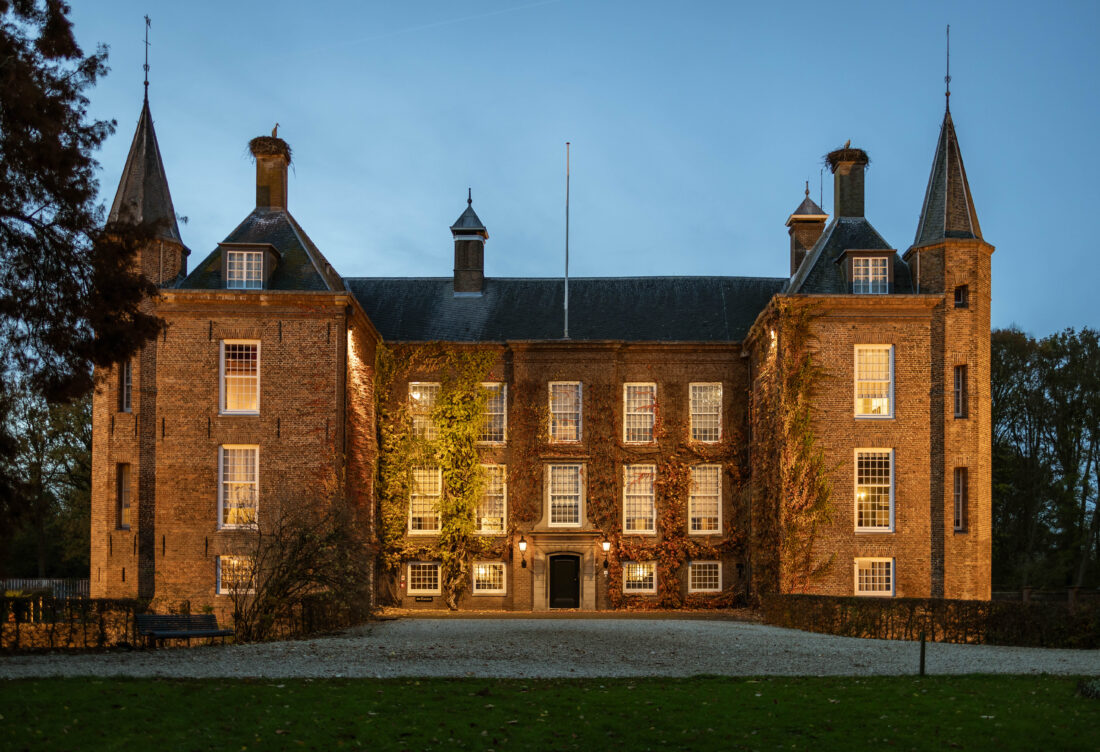 Front facade of Slot Zuylen castle at dusk with brick architecture, symmetric windows, conical towers, and illuminated ground against a twilight sky