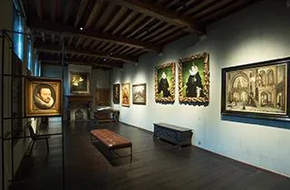 Sala de galería de museo con techo de vigas de madera a la vista, suelos de madera oscura, bancos de madera y cuadros enmarcados colgados en las paredes.