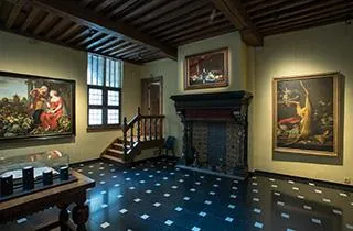 Habitación interior histórica con suelo de damero en blanco y negro, vigas de madera oscura en el techo, chimenea, escalera de madera y grandes cuadros enmarcados colgados en paredes de color crema.