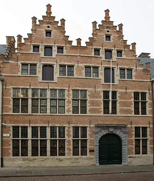 Fachada de ladrillo de un edificio flamenco histórico con dos hastiales escalonados, varias filas de ventanas y una gran puerta arqueada de color verde.