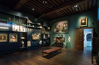 Sala de museo con paredes de color verde oscuro, techo con vigas de madera, cuadros enmarcados, vitrinas de cristal y un banco de madera en el centro