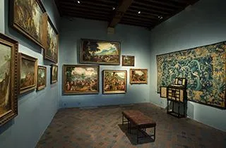 Sala de museo con cuadros enmarcados en paredes azules y un gran tapiz, con un banco de madera en el centro y un soporte informativo.
