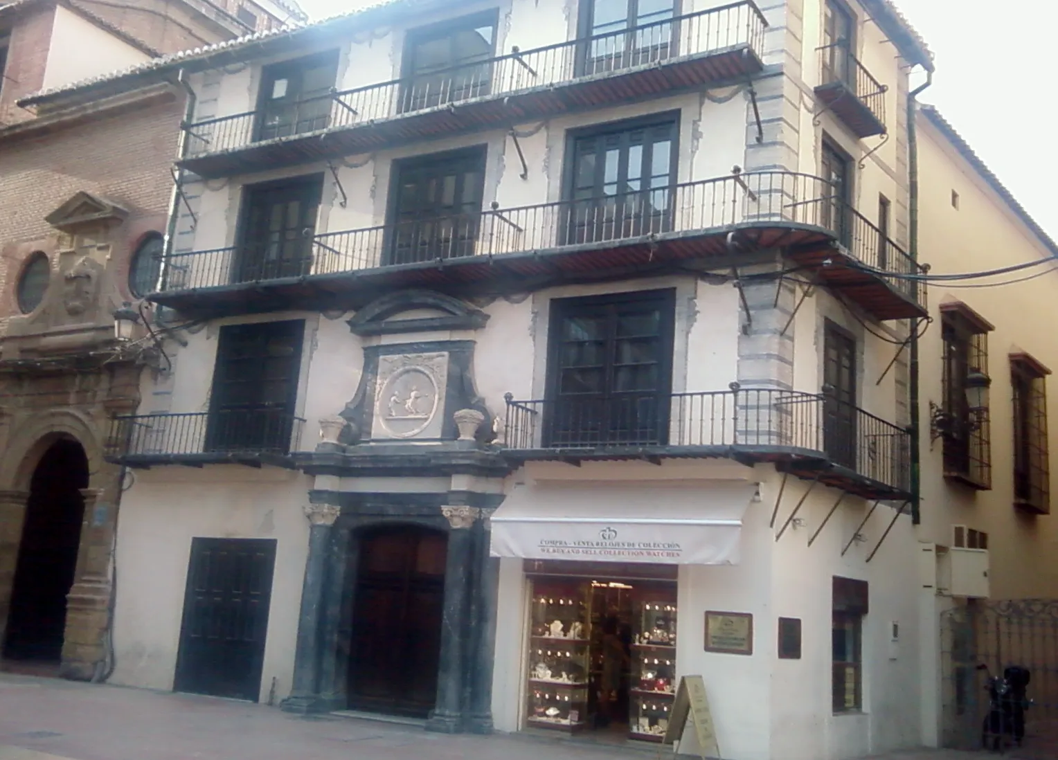 Vista a pie de calle de la fachada de la Casa del Consulado en Málaga, un edificio histórico blanco con balcones de hierro negro, puertas y ventanas de madera oscura, y una entrada a una tienda en la planta baja.