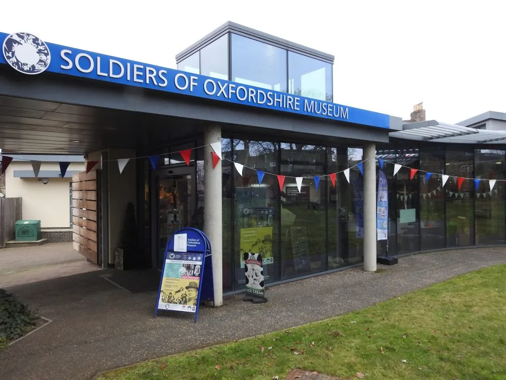 Vista exterior de la entrada del Soldiers of Oxfordshire Museum con señalización azul, fachada de cristal y banderas de colores