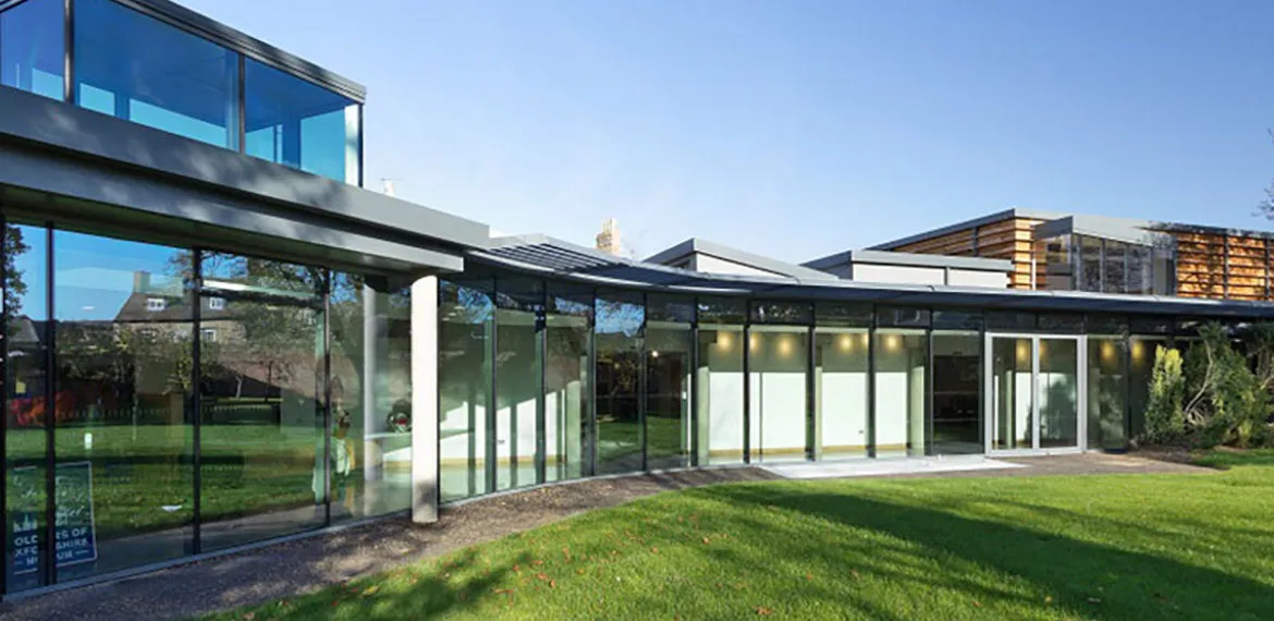 Exterior de un edificio de museo moderno con grandes muros cortina de cristal, voladizo de techo plano, columnas de acero y un césped verde bajo un cielo azul despejado.